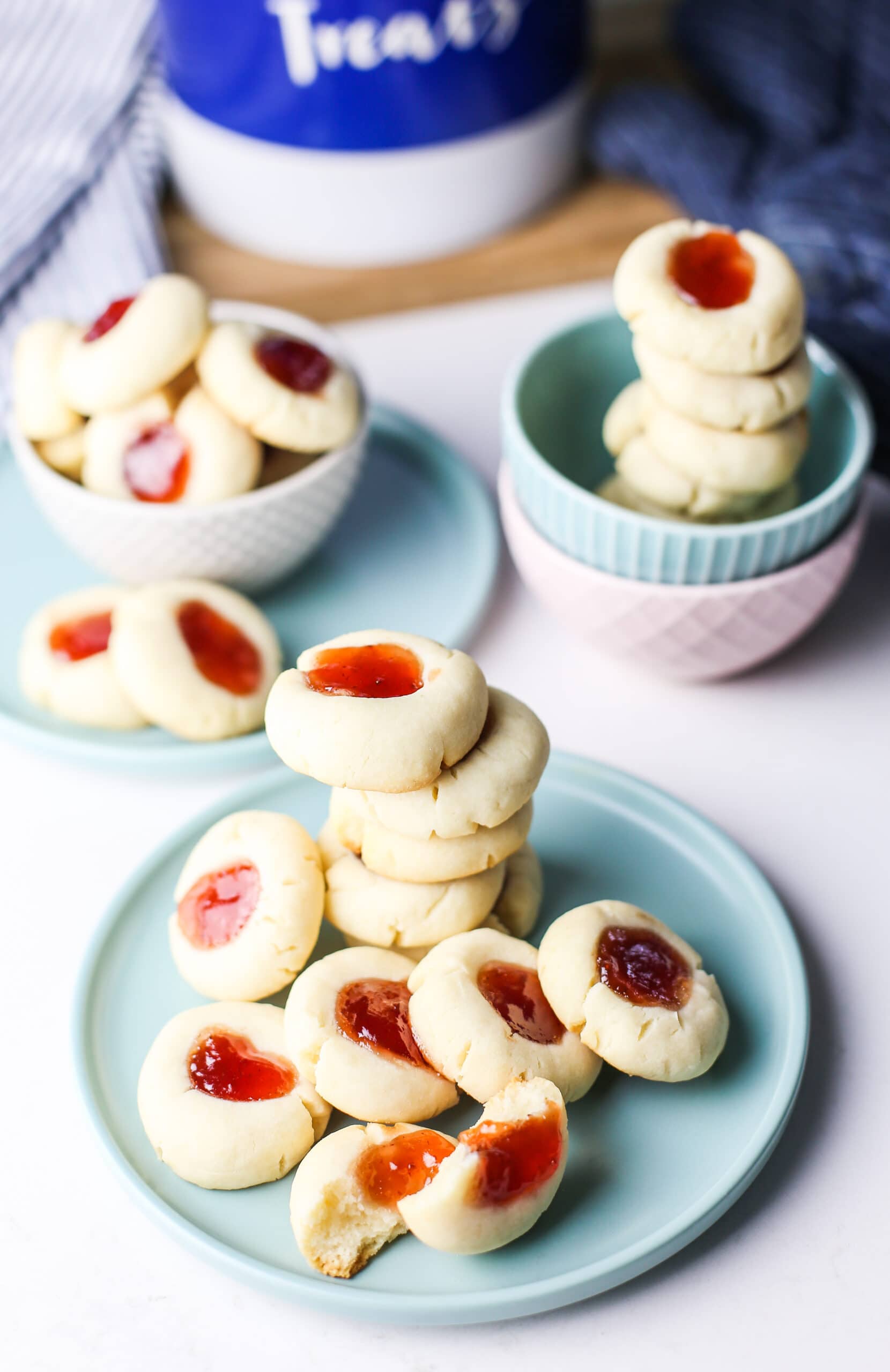 A top angled view of condensed milk thumbprint jam cookies on a blue plate and in two bowls.