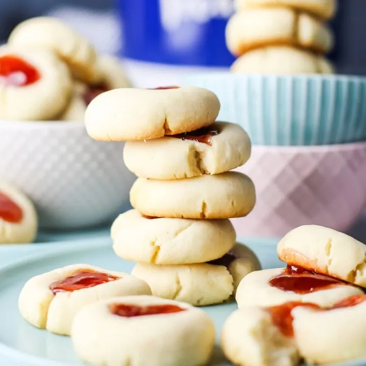Five stacked Condensed Milk Thumbprint Cookies on a blue plate surrounded by more cookies.