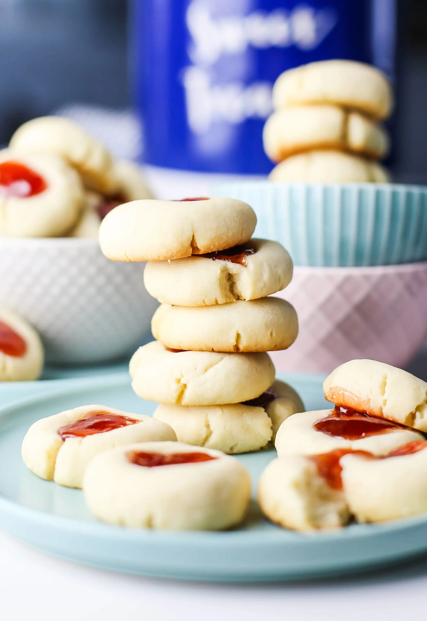 Five stacked Condensed Milk Thumbprint Cookies on a blue plate surrounded by more cookies.