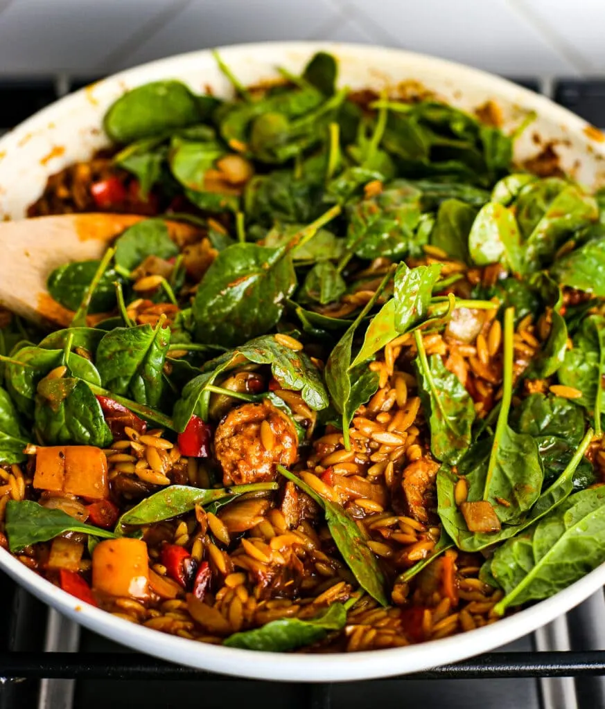 Fresh baby spinach being stirred into a white pan with sliced Italian sausage, cooked orzo, and vegetables in a red sauce.