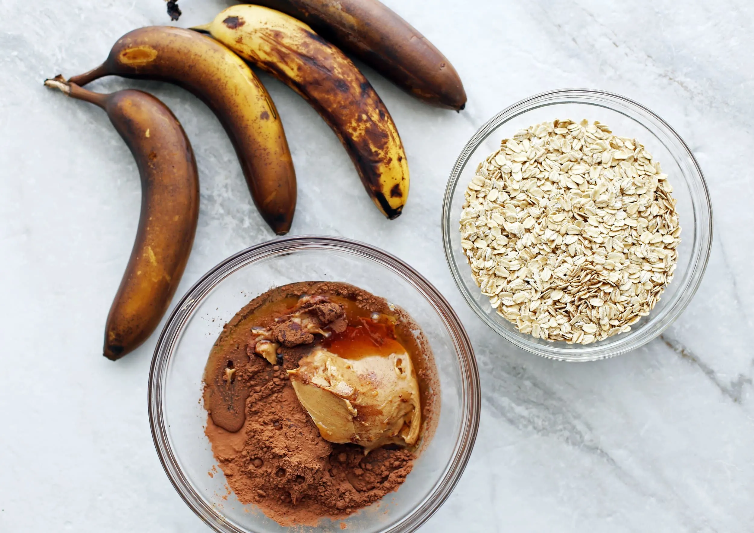 Four ripe banana, a bowl of peanut butter, cocoa powder, and maple syrup, and another bowl with oats.