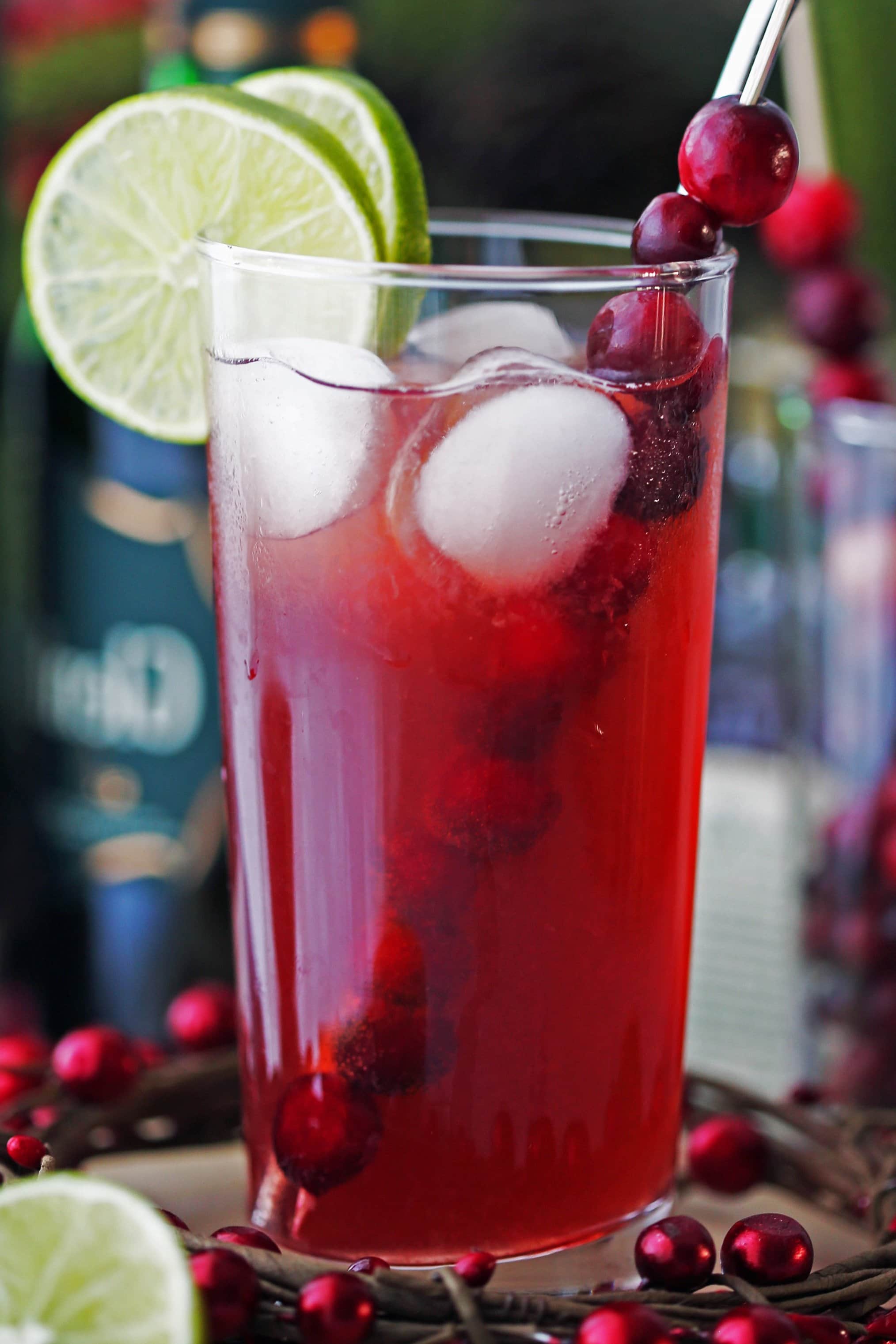 A tall highball glass of Cranberry Whiskey Ginger Cocktail with lime slices, fresh cranberries, and ice cubes.