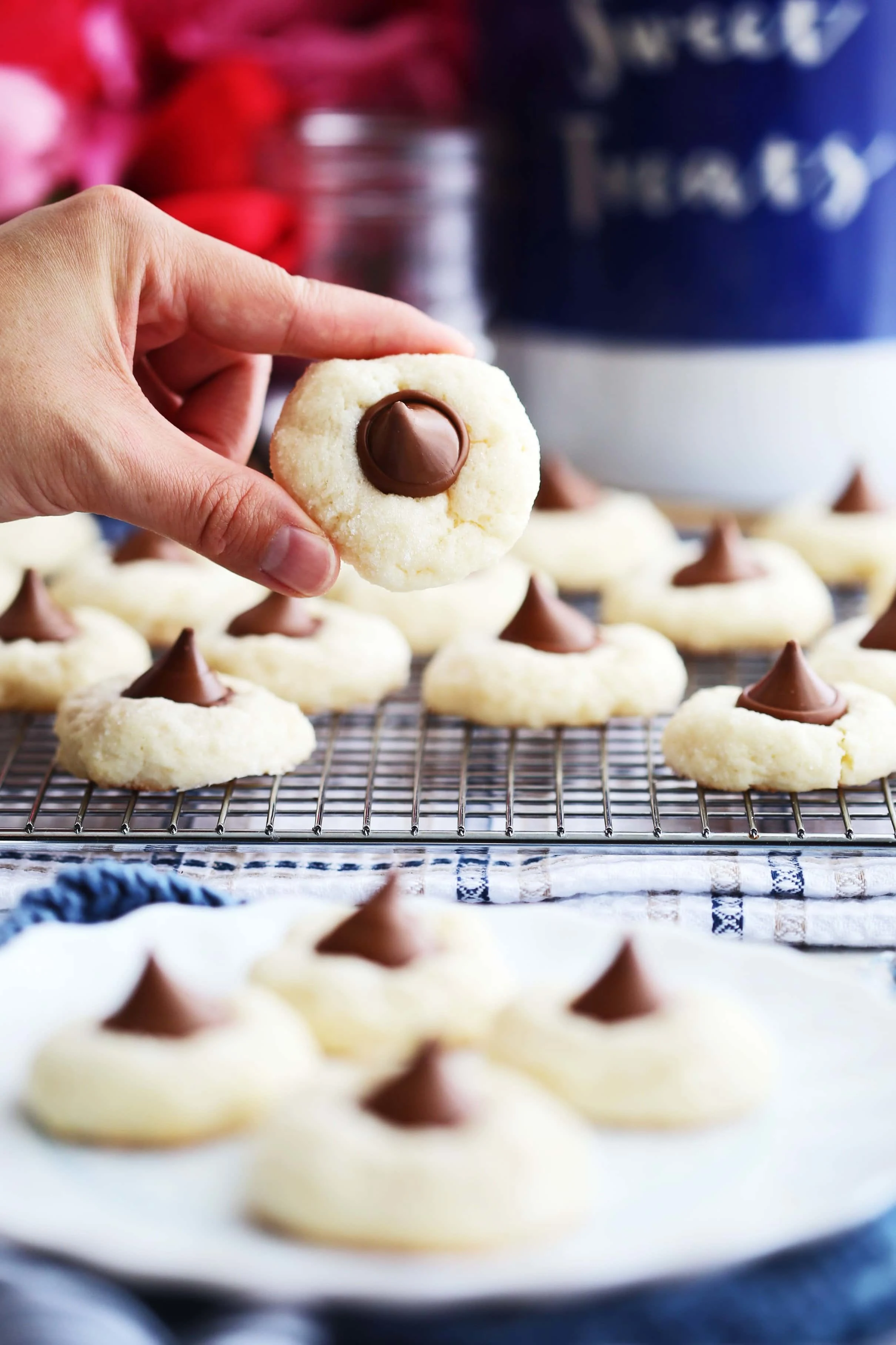 A hand holding up a single Cream Cheese Kiss Cookie over a white plate and cooling rack filled with more Cream Cheese Kiss Cookies.