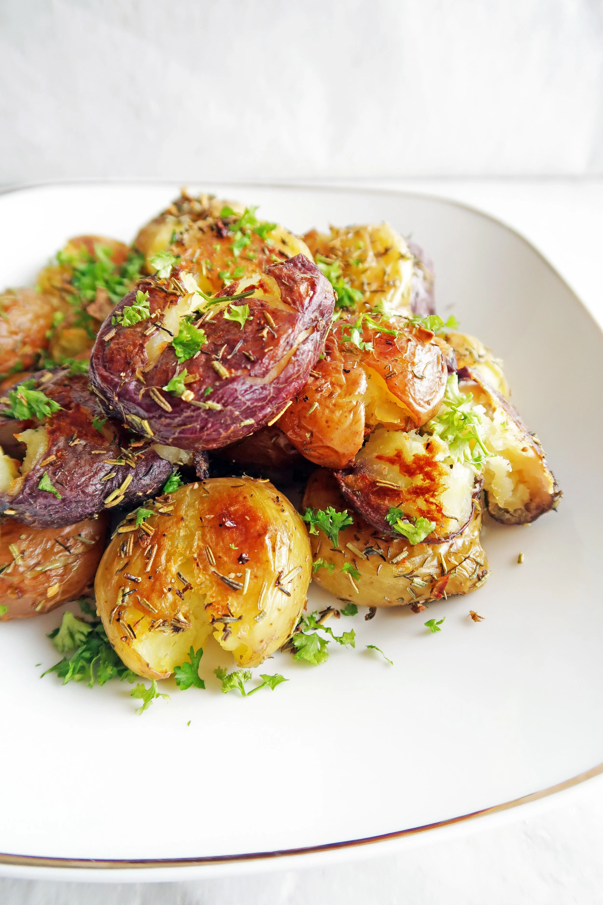 A side view of vegan and gluten-free Crispy Garlic Smashed Baby Potatoes piled on a white plate and topped with parsley.