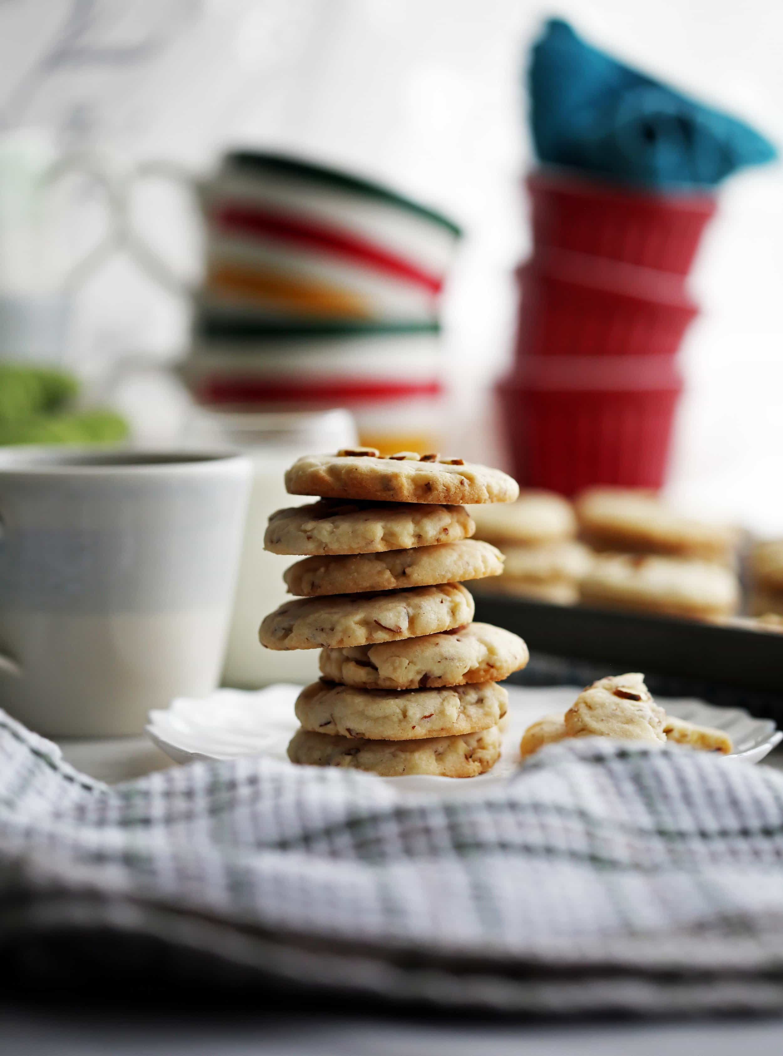 A stack of seven crunchy almond cookies on a white plate with cookies and coffee in the background.