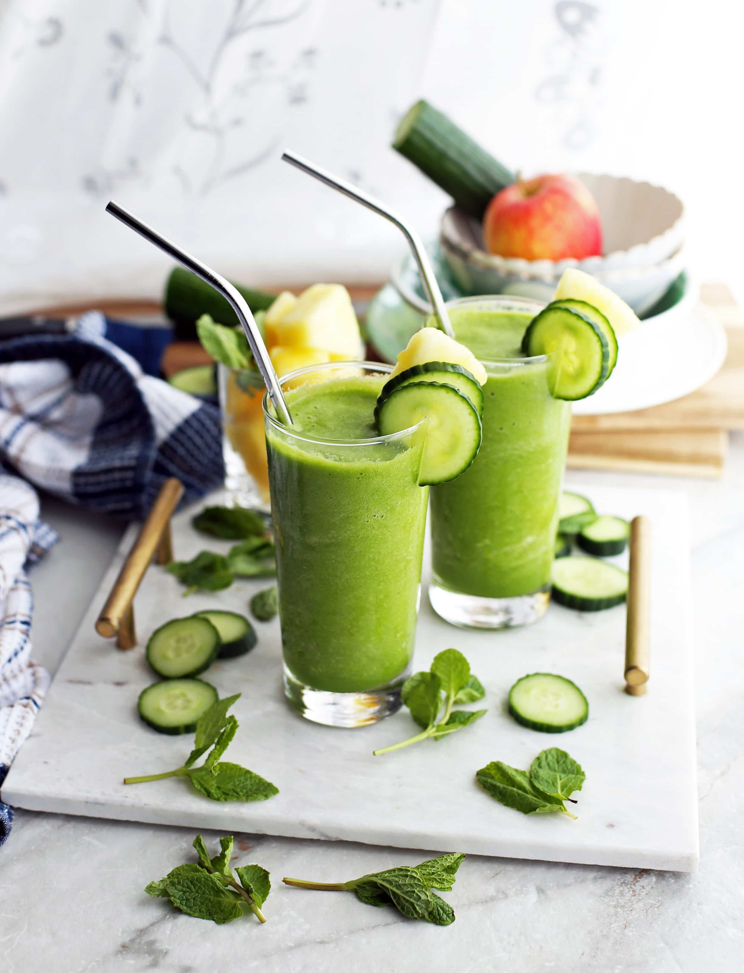 Two glasses of Cucumber Mint Pineapple Smoothies sitting on a marble platter and surrounded by fresh mint and cucumber slices.