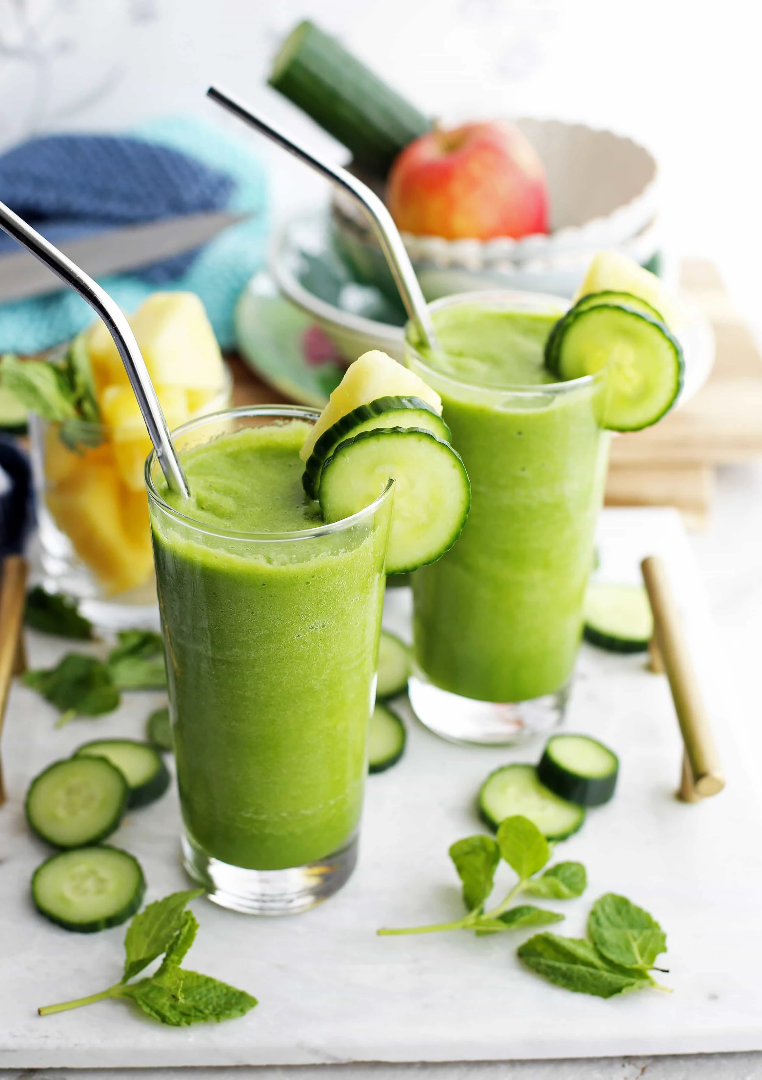 Two Cucumber Mint Pineapple Smoothies in tall glasses that are garnished with cucumber slices, pineapple slices, and straws.