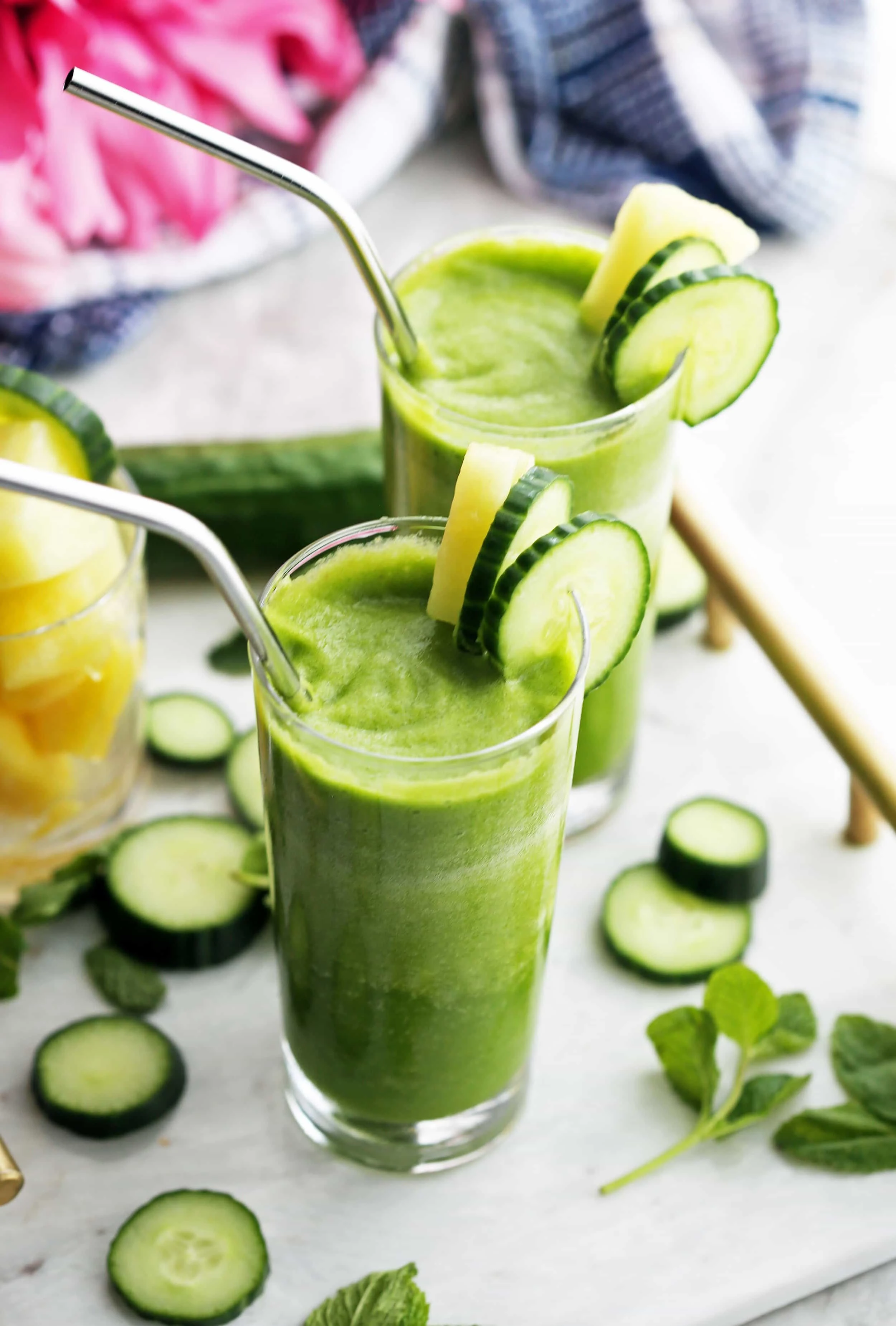 Two tall glasses of Cucumber Mint Pineapple Green Smoothies garnished with and surrounded by cucumber and pineapple slices and fresh mint.