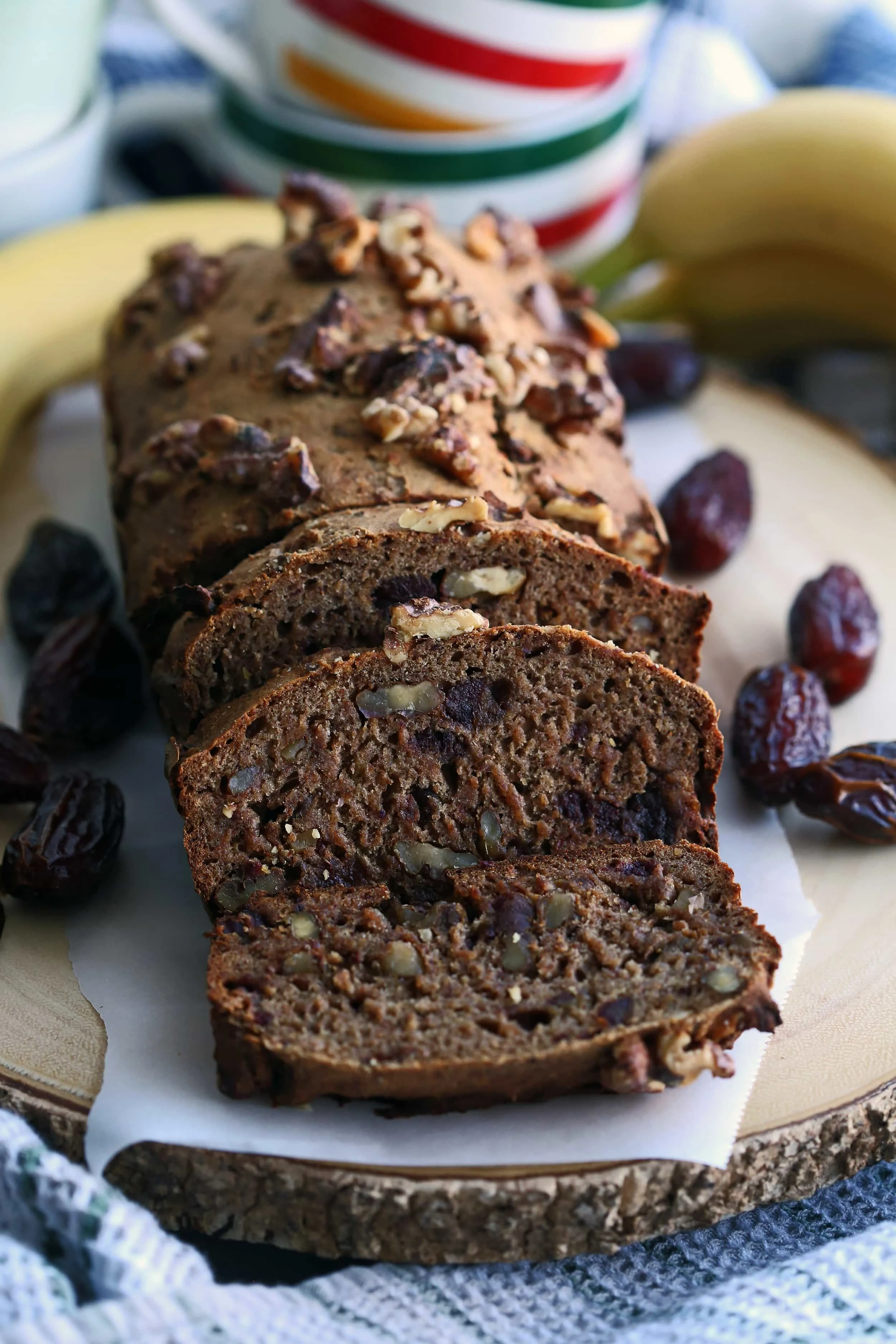 A loaf of banana bread with Medjool dates and walnuts surrounded by fresh bananas and dates on a wooden platter.