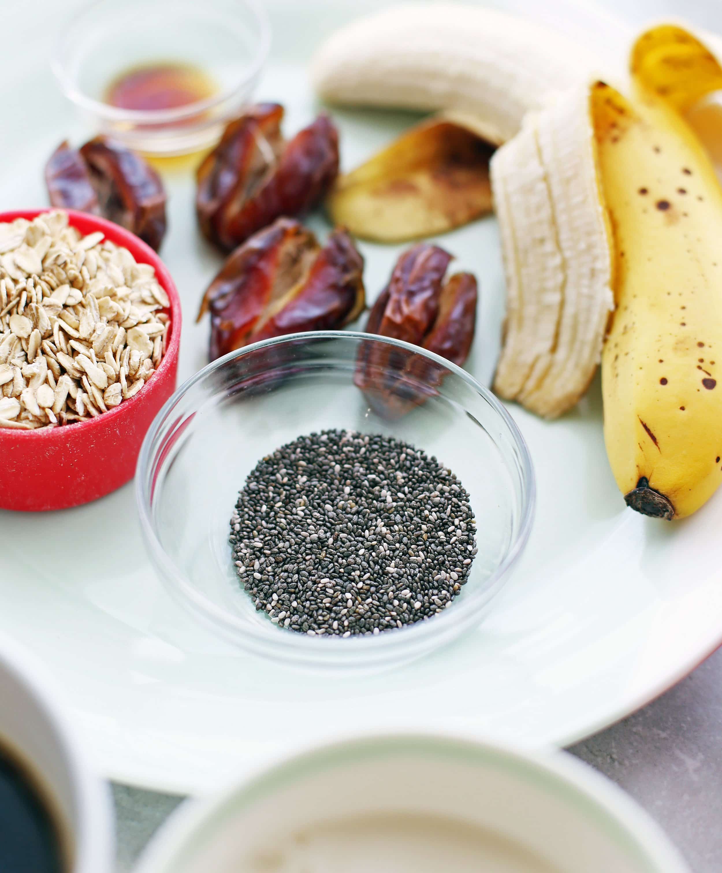 A closeup of a small bowl containing chia seeds; dates, banana, rolled oats, and vanilla extract in the background.