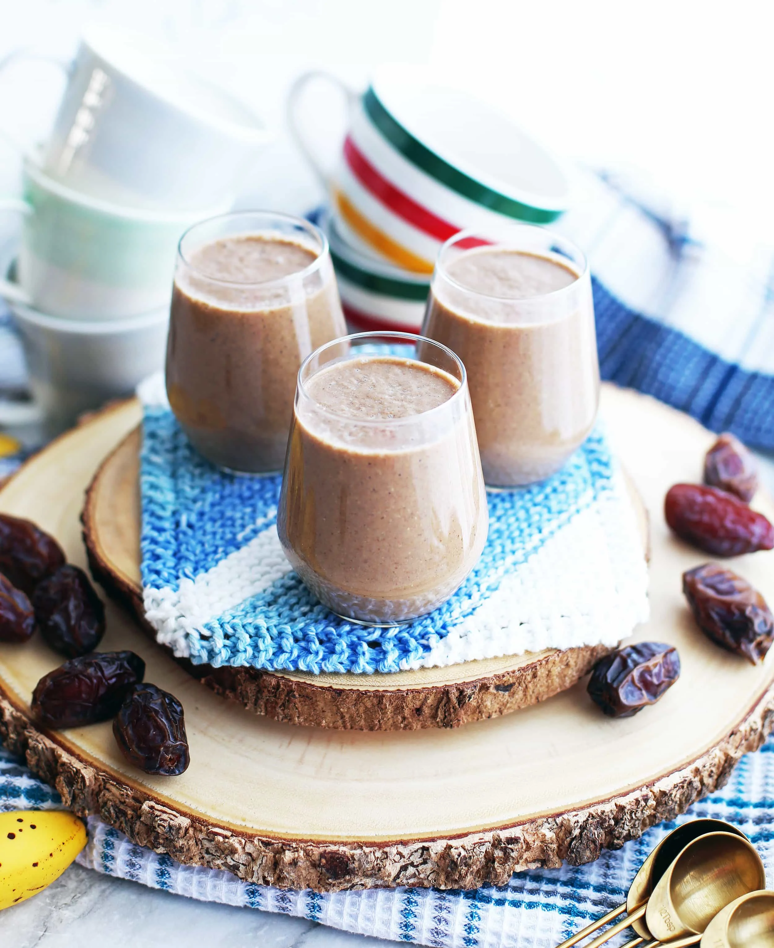 Three glasses of creamy date coffee breakfast smoothies on a wooden platter surrounded by medjool dates and coffee mugs.
