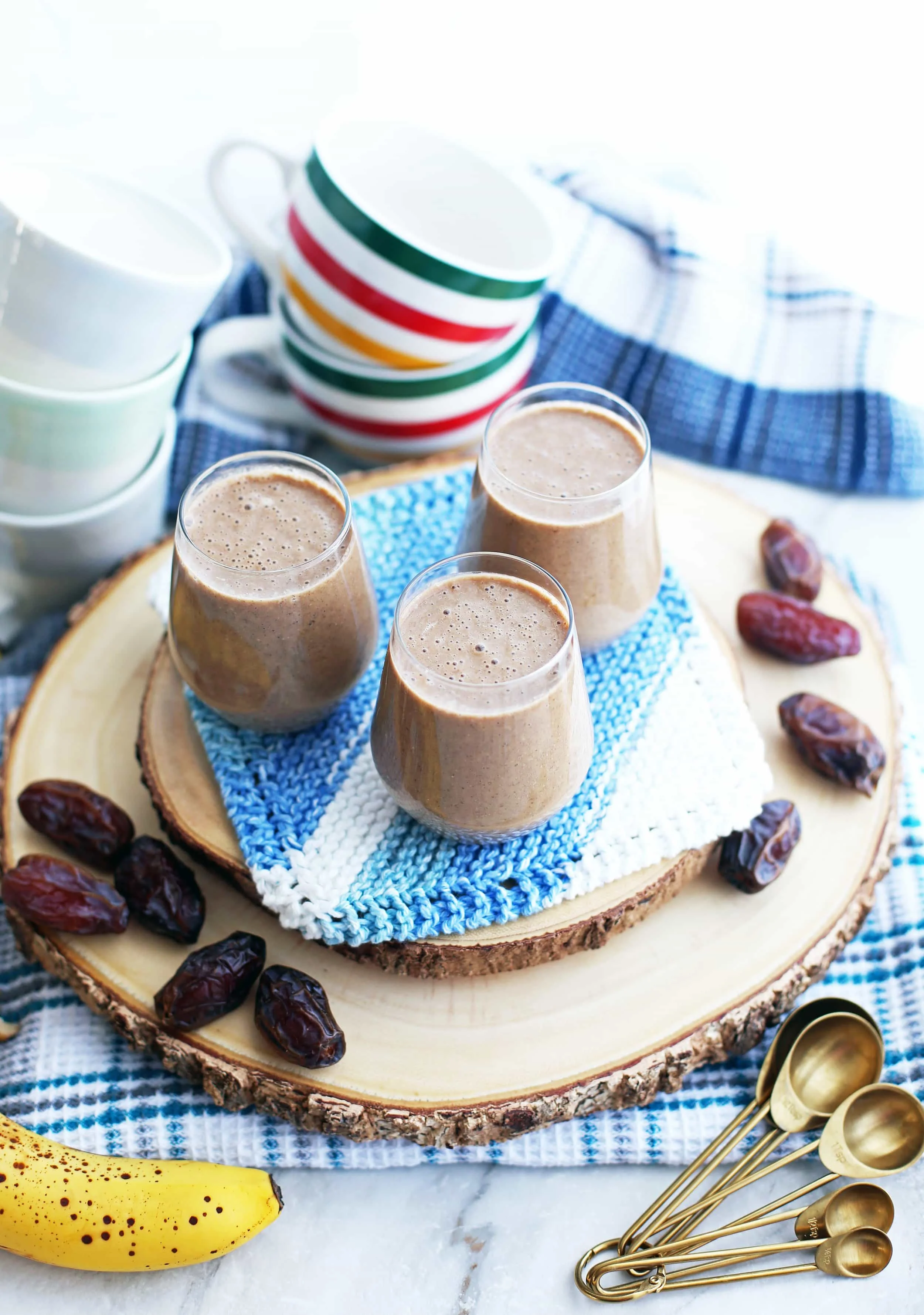 Three glasses of creamy date coffee breakfast smoothies on a wooden platter surrounded by medjool dates, measuring spoons, and coffee mugs.