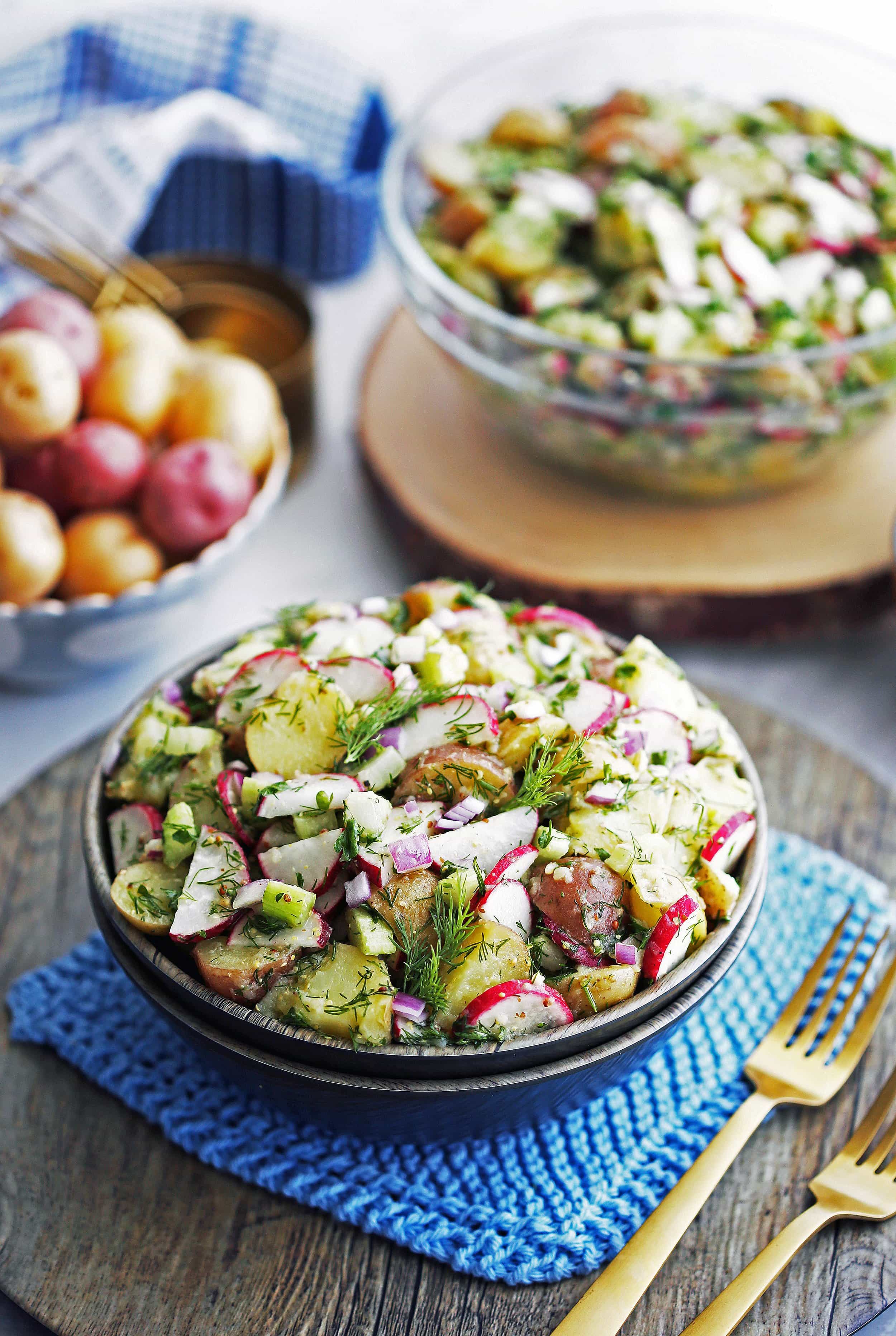 A wooden bowl full of Mustard Dill Potato Salad with more salad in a glass bowl, and small bowl of baby potatoes behind it.