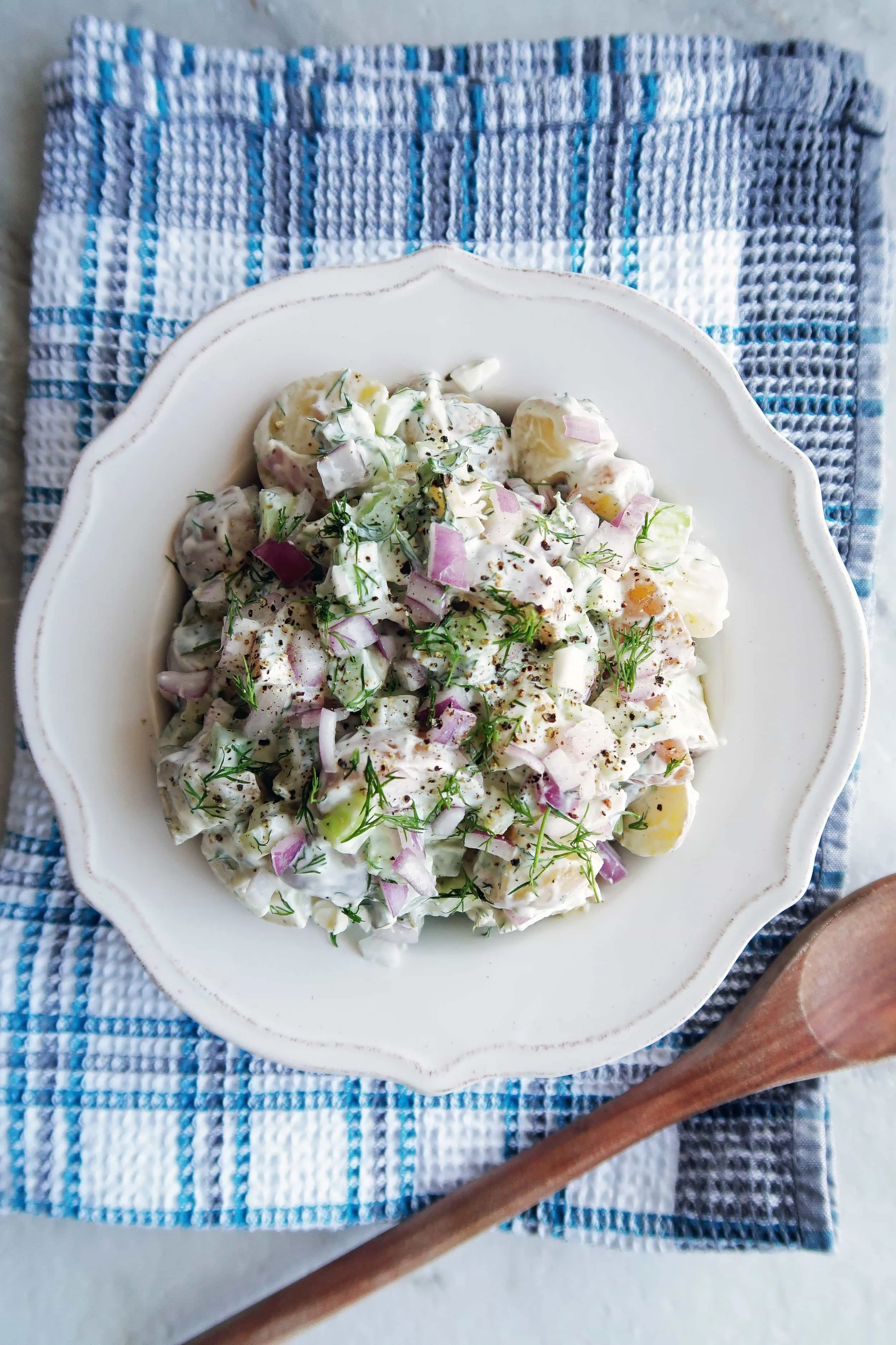 A large white bowl of Dill Pickle Potato Salad on a blue checkered towel and wooden spoon to the side.