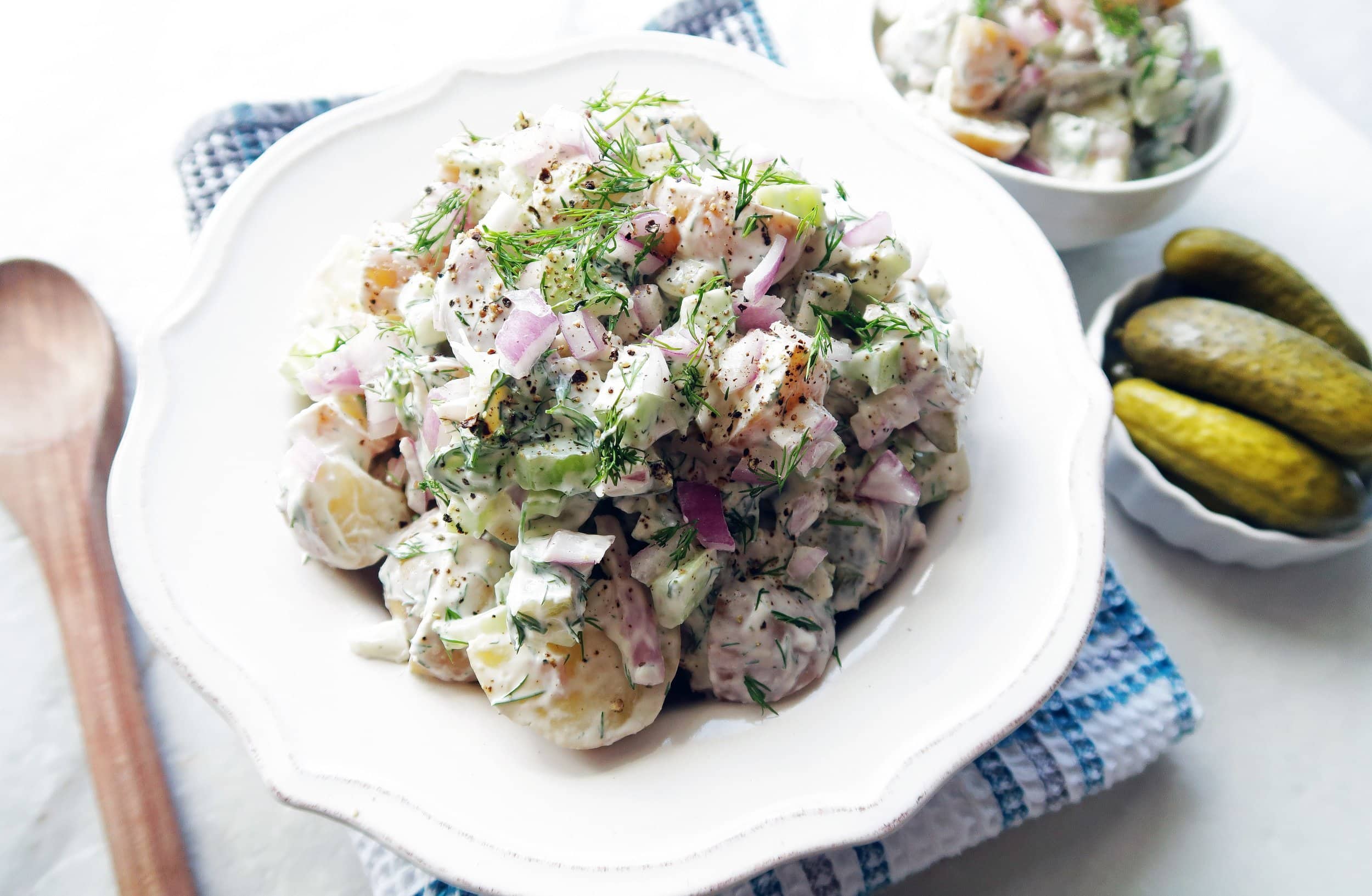 A white bowl of Dill Pickle Potato Salad with wooden spoon, pickles, small bowl of salad around it.