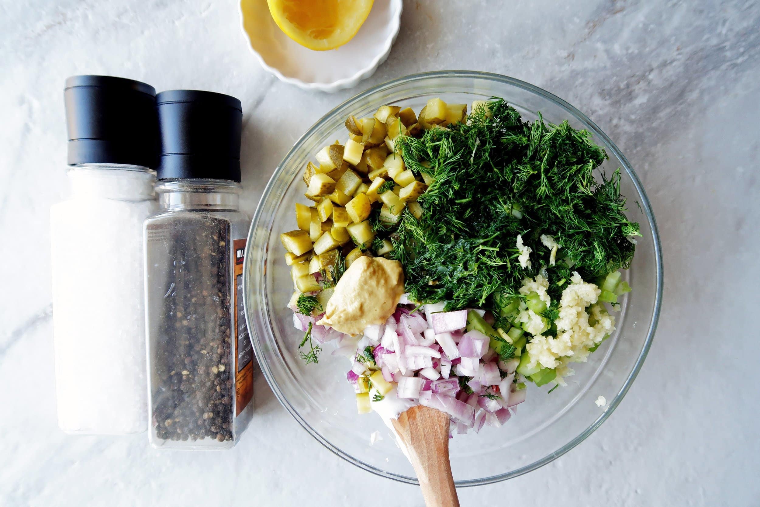 Chopped potatoes, onions, pickles, dill, garlic, and celery in a large glass bowl; salt and pepper mills on the side.