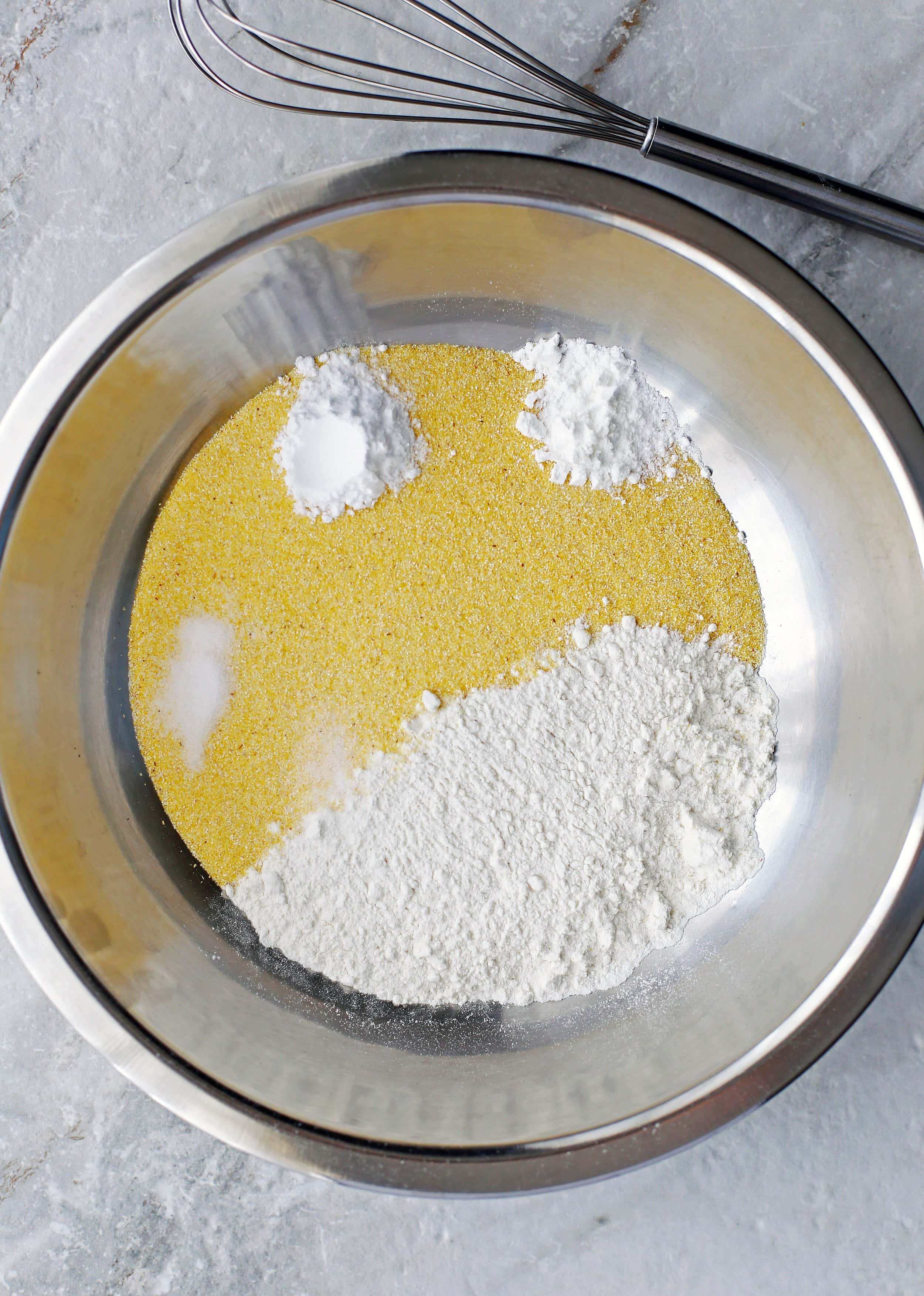 A large metal bowl containing cornmeal, flour, baking soda, baking powder, and spices.