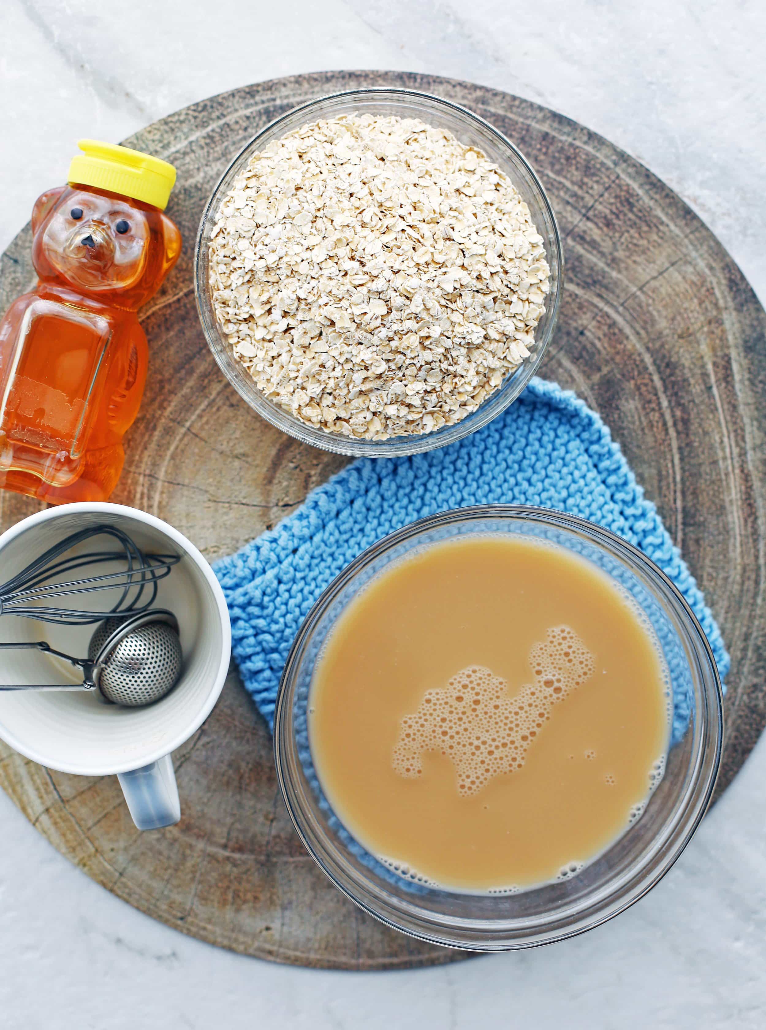 A bowl of Earl Grey Tea, honey, rolled oats, coffee cup, and metal whisk on a round wooden platter.
