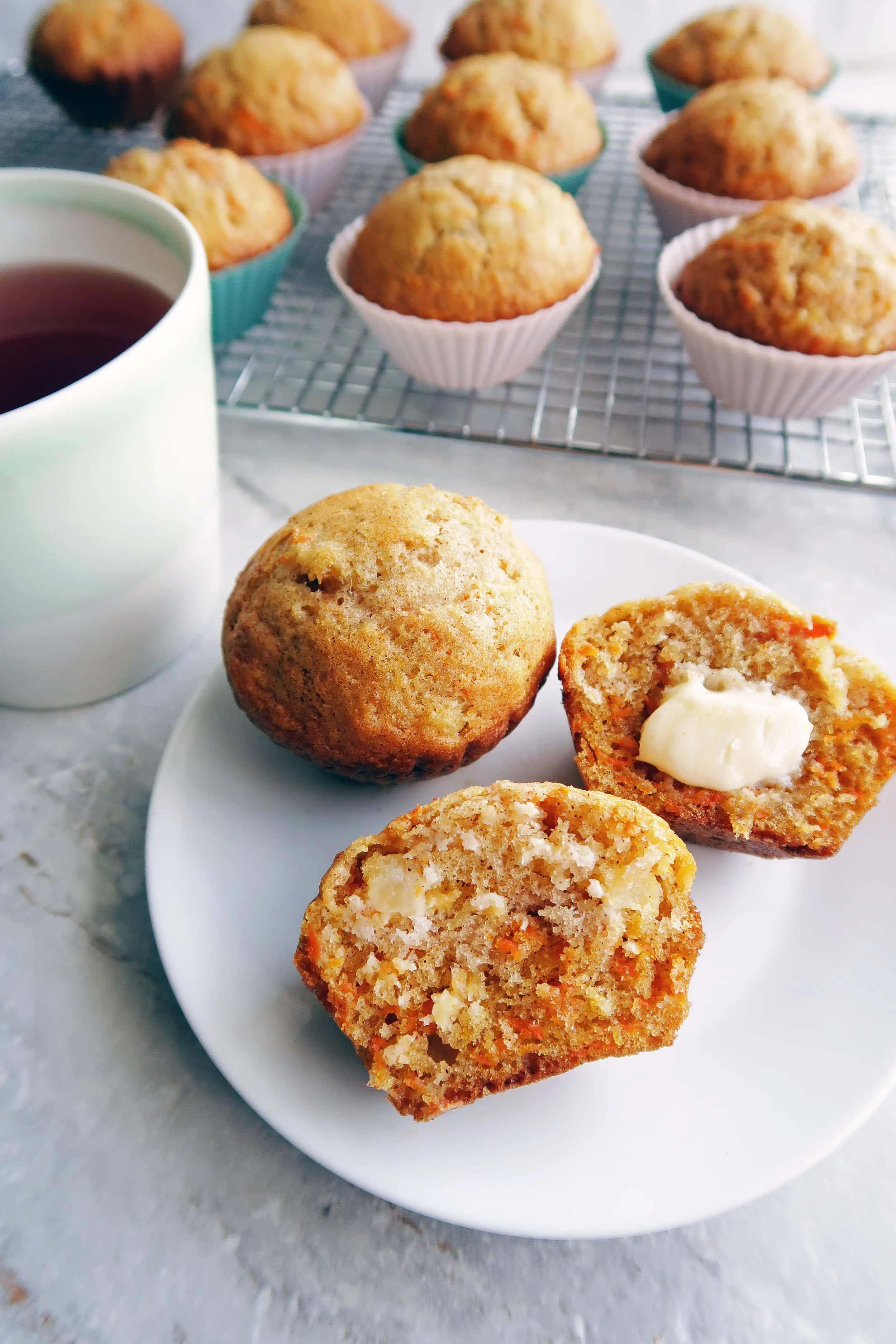 Two Carrot Pineapple Muffin, one cut in half with butter, on a white plate with more muffins on a cooling rack in the background.