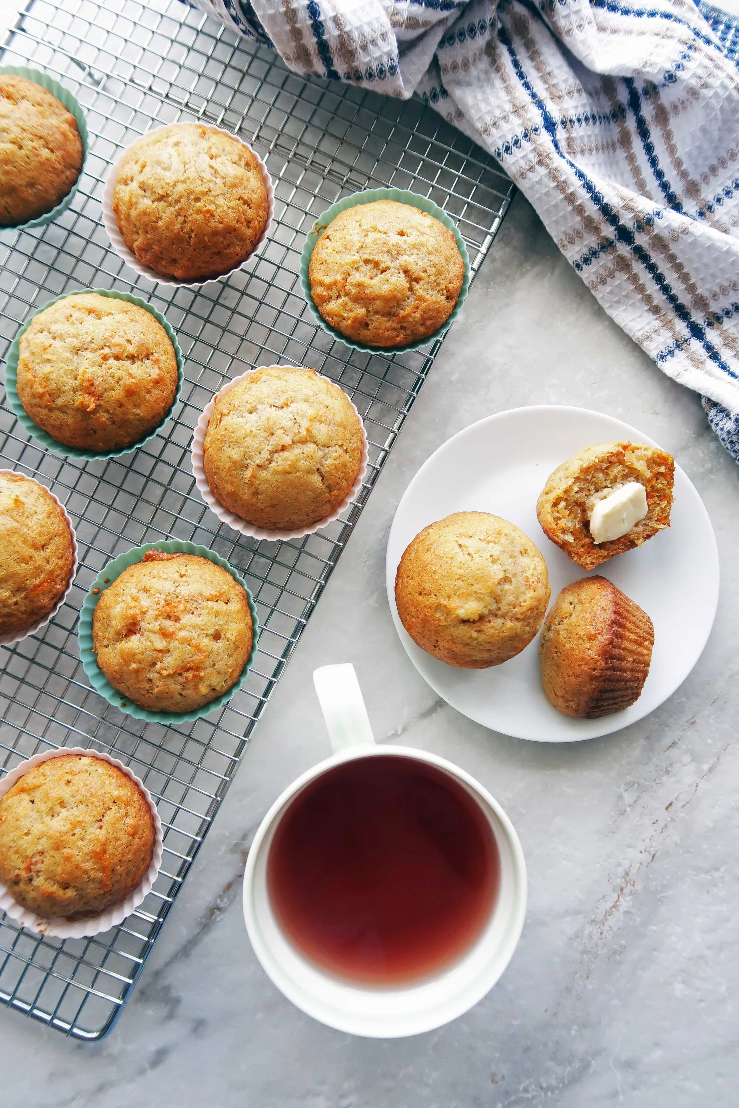 Overhead view of two carrot Pineapple Muffins on small white plate, muffins on a cooling rack, and a cup of tea.