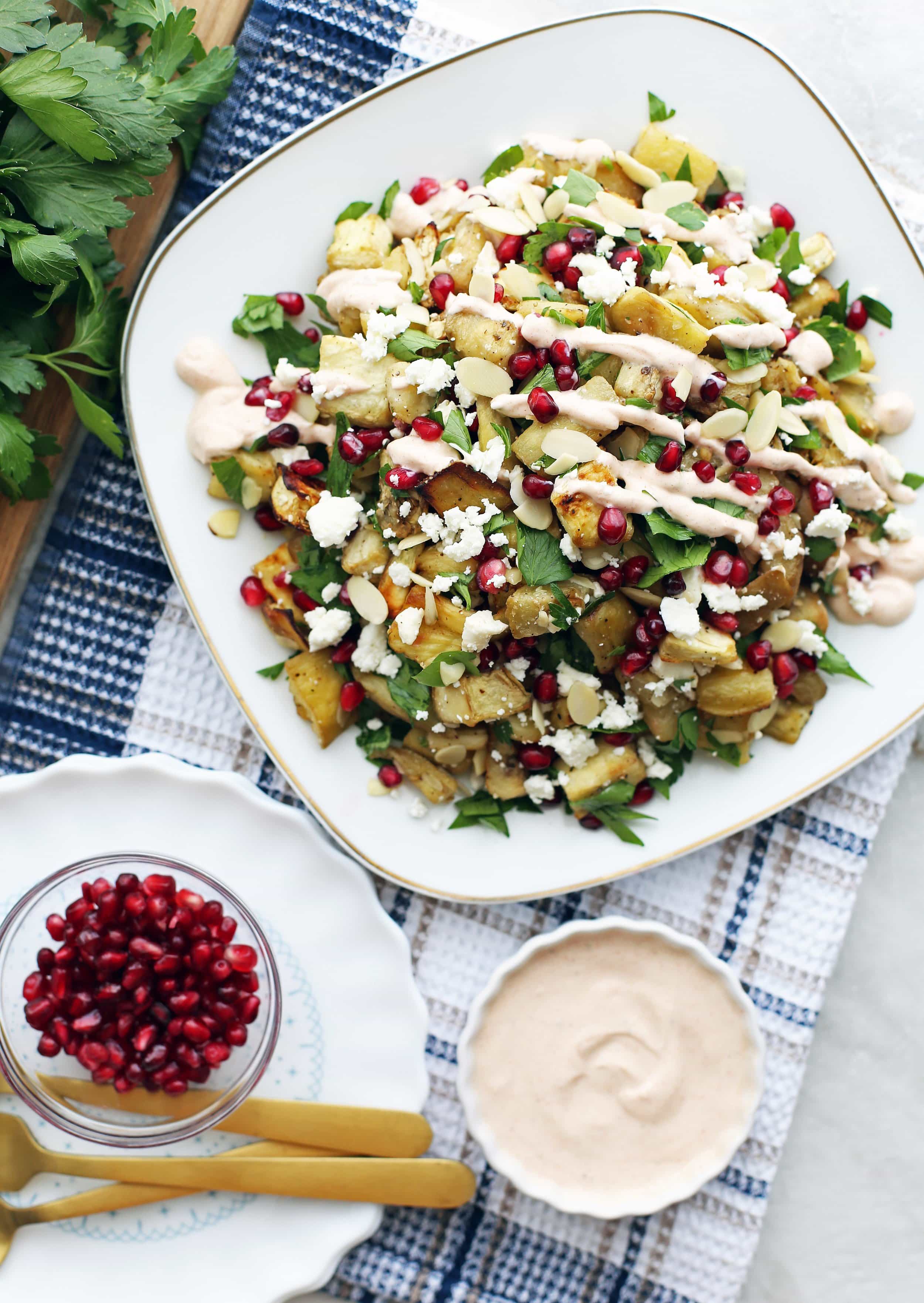 Roasted eggplant pomegranate feta salad on a white plate,and smoked paprika yogurt dressing and pomegranate seeds in two small bowls.