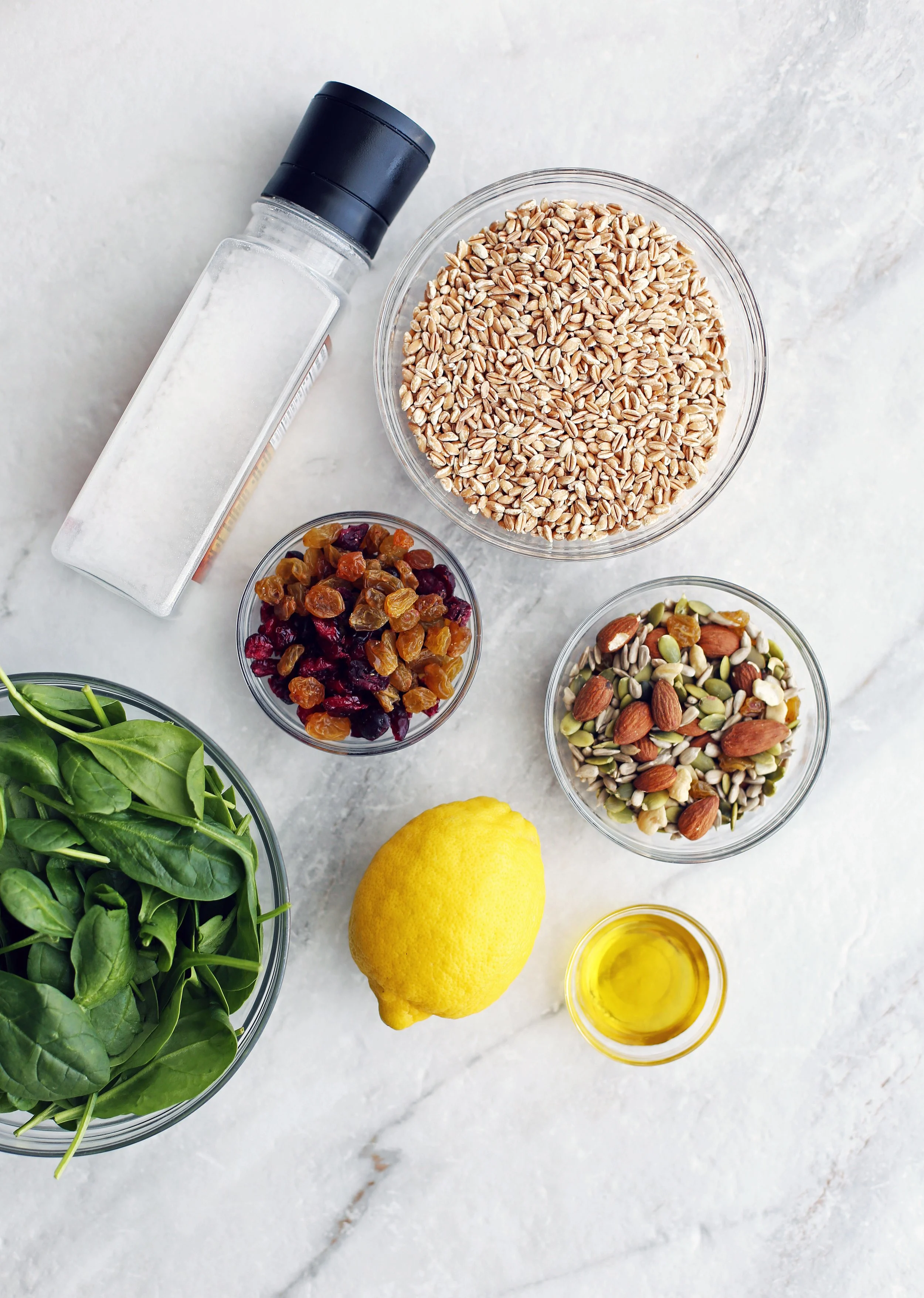 Bowls of baby spinach, dried fruit, nuts and seeds, farro, olive oil along with a lemon and a salt shaker.