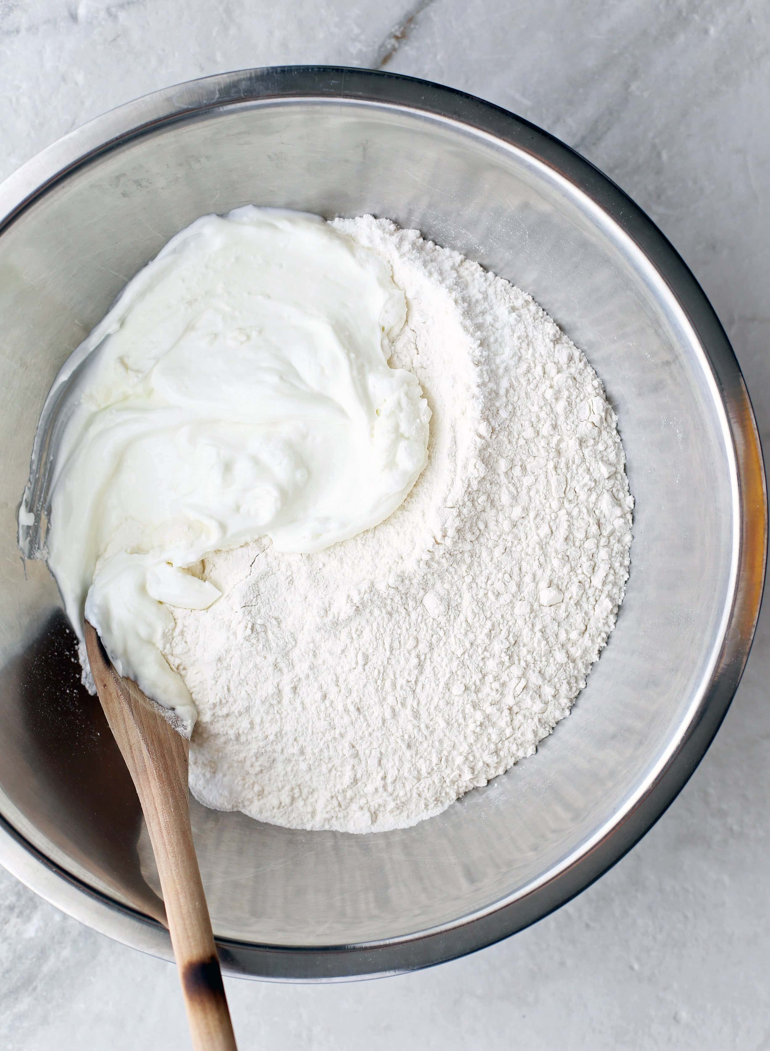 Greek yogurt, all-purpose flour, and baking powder in a large metal bowl with a wooden spoon.