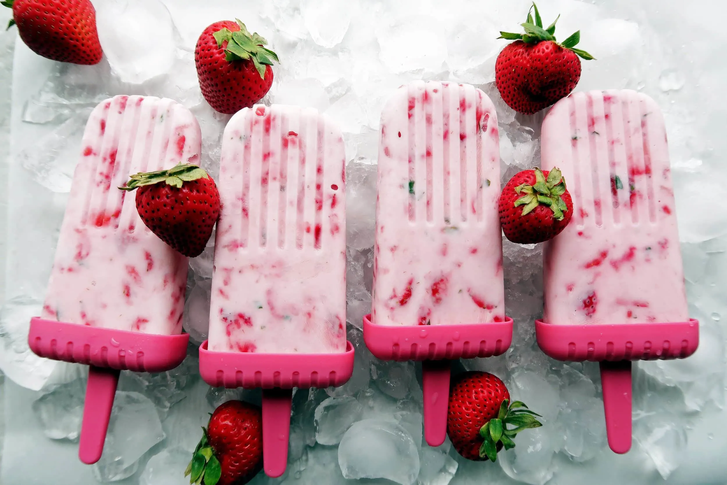 Four Fresh Strawberry Mint Yogurt Popsicles placed side by side on a bed of ice with strawberries around it.