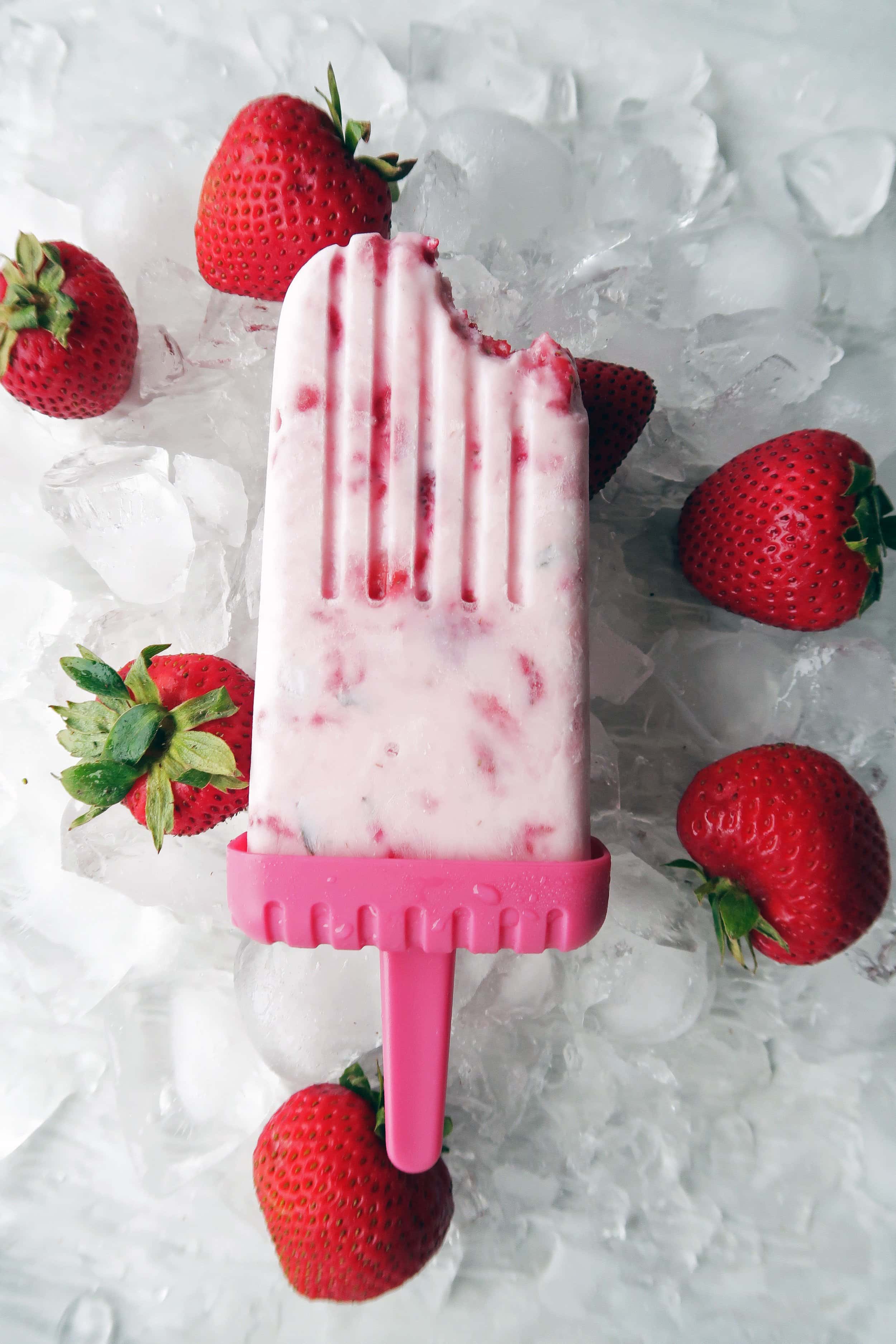 A Fresh Strawberry Mint Yogurt Popsicle with a bite taken that placed on a bed of ice with strawberries around it.