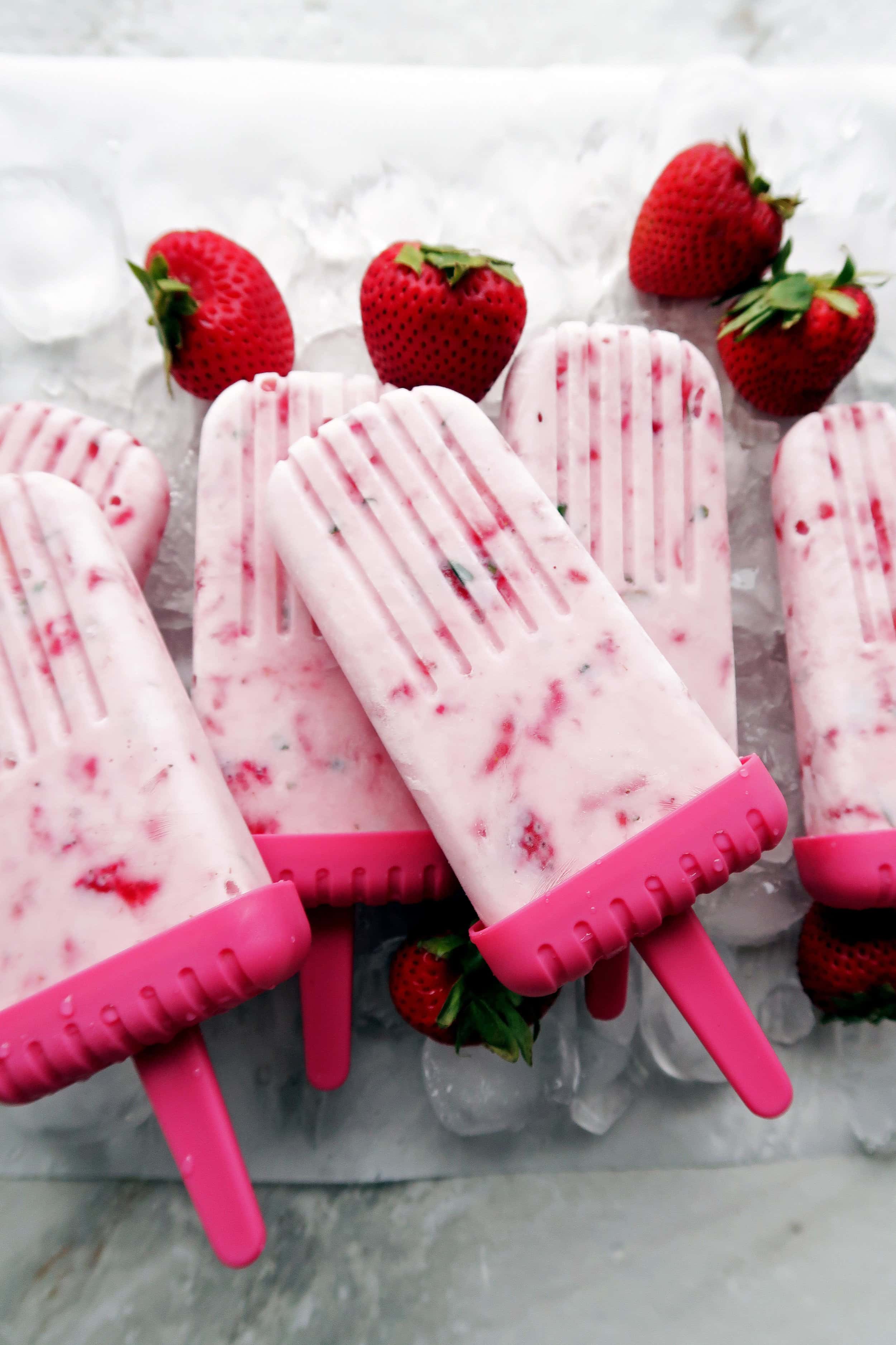 Six Fresh Strawberry Mint Yogurt Popsicles placed on top of one another on a bed of ice with strawberries around it.