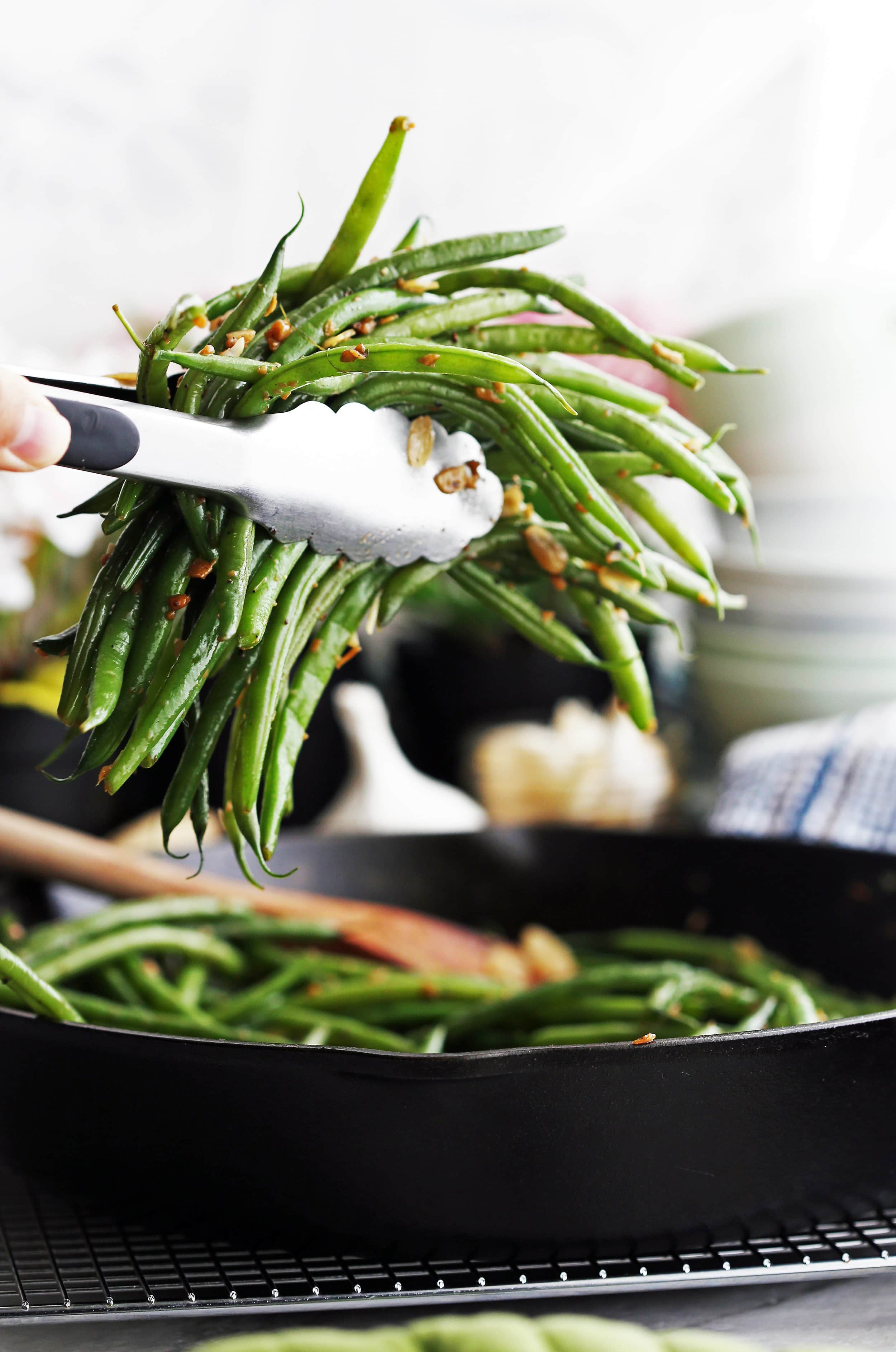 Metal tongs holding garlic ginger green beans with more green beans in a cast iron skillet below.