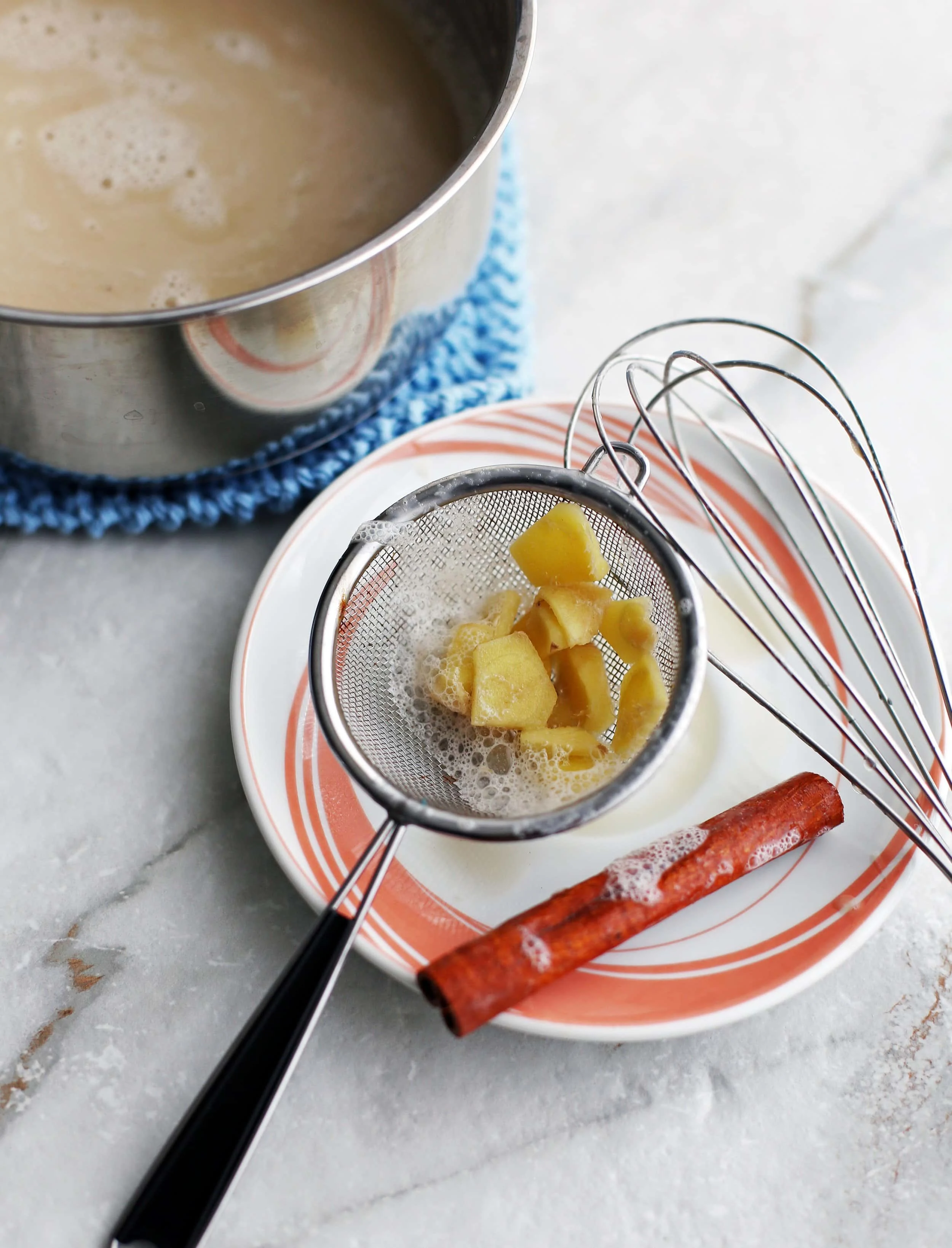 A angled top view of sliced ginger in a small strainer, one cinnamon stick, and a whisk.