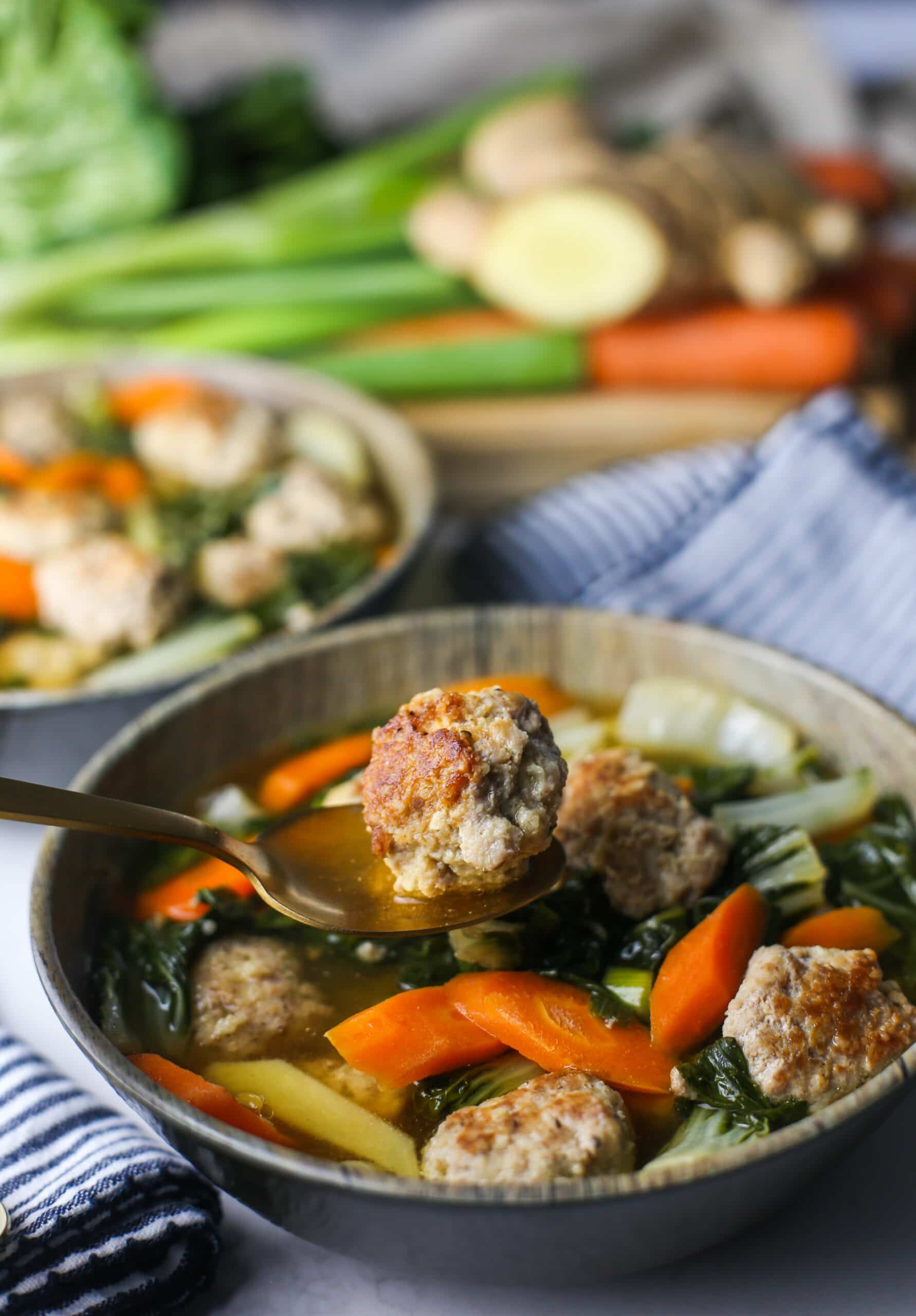 A gold spoon holding up a single ginger pork meatball over bowl of pork meatball soup with bok choy.