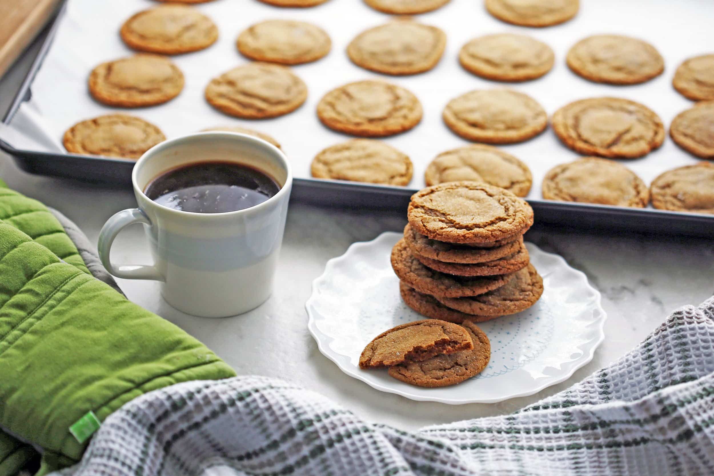 A side view of a plate of chewy gingersnap cookies with a cup of coffee and cookies on a baking sheet in the background.