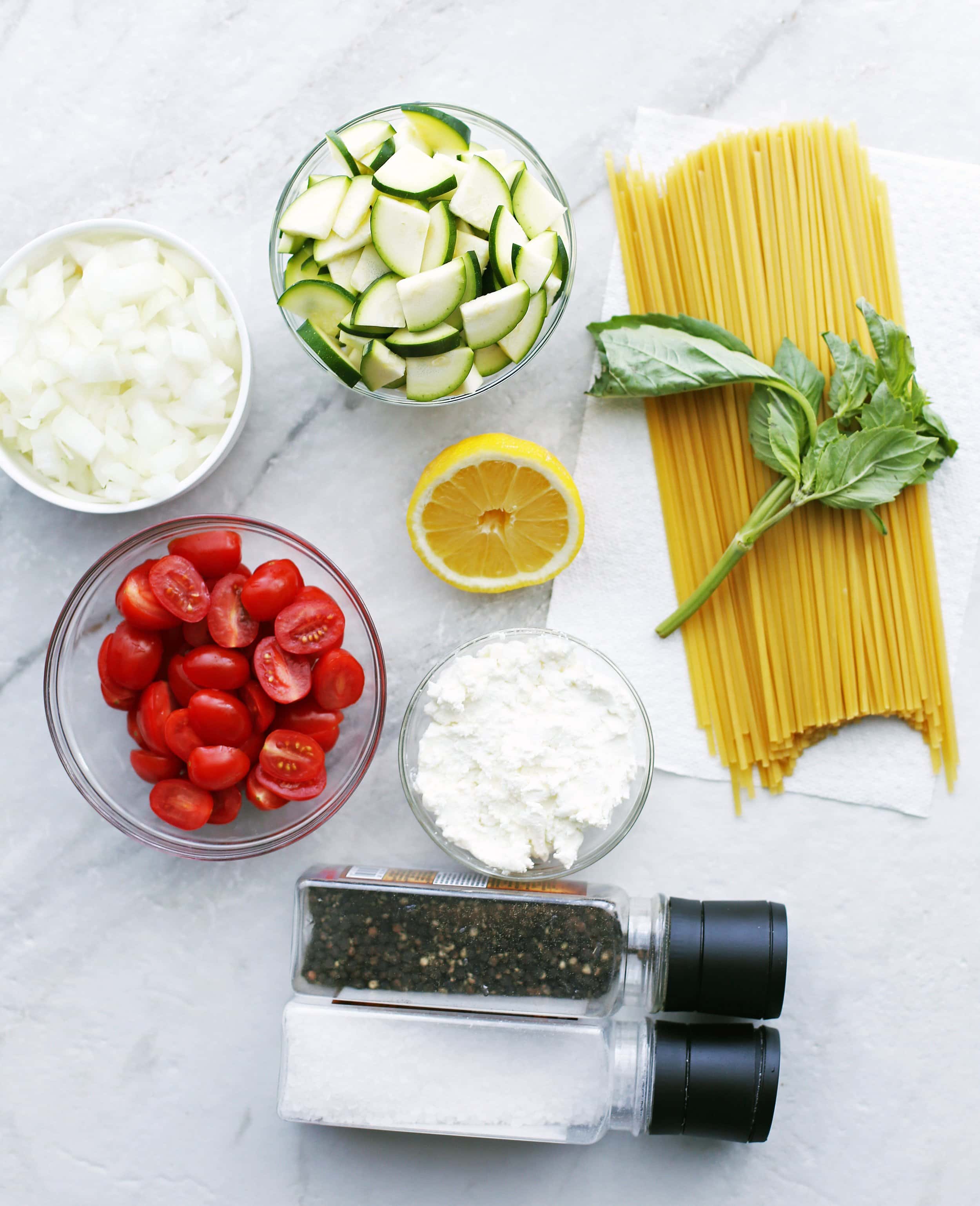 Bowls of grape tomatoes, chopped onion, chopped zucchini, lemon, goat cheese, dry linguine, fresh basil, and salt and pepper shakers.