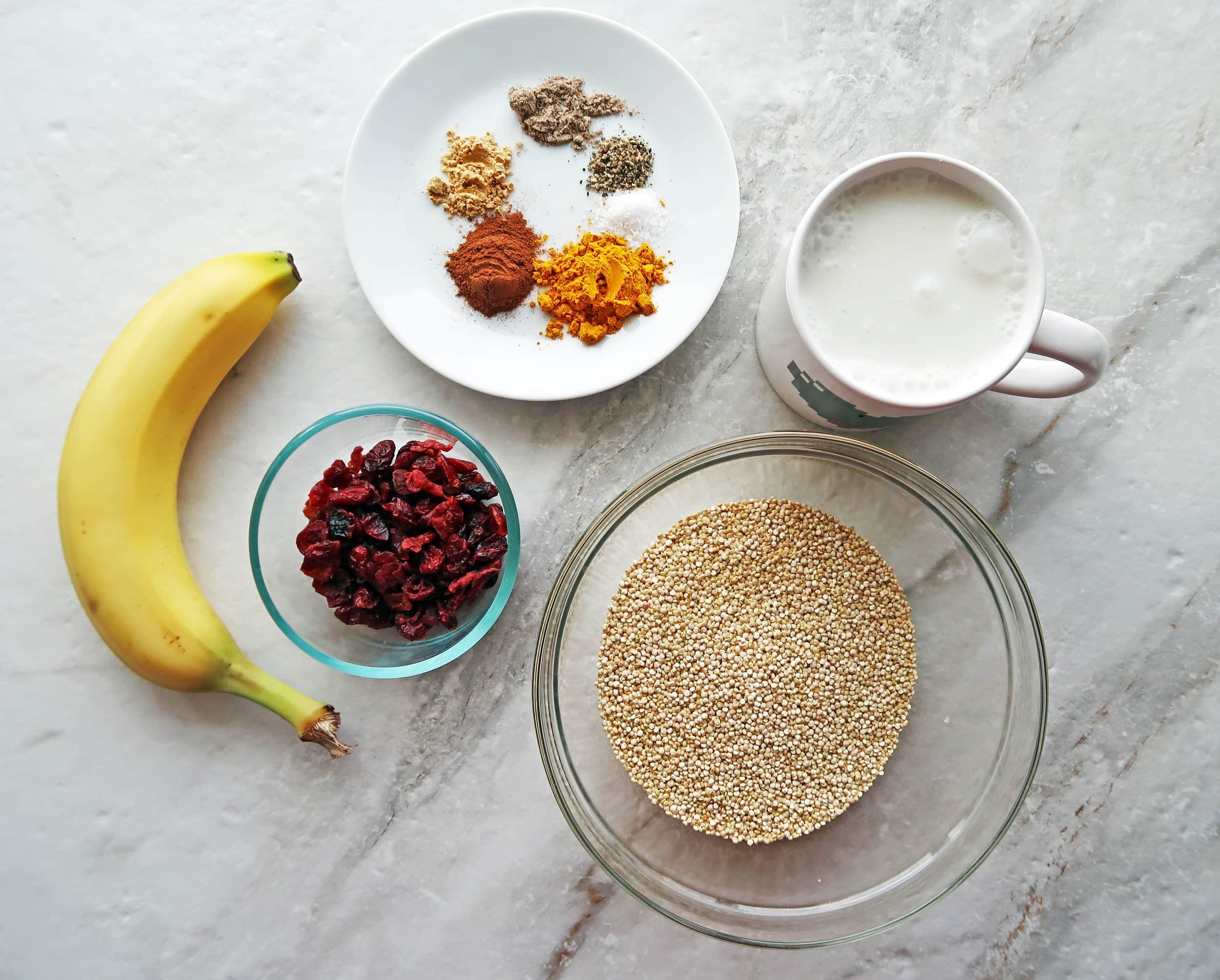 Bowls of uncooked quinoa, dried cranberries, spices, coconut milk, and a banana.