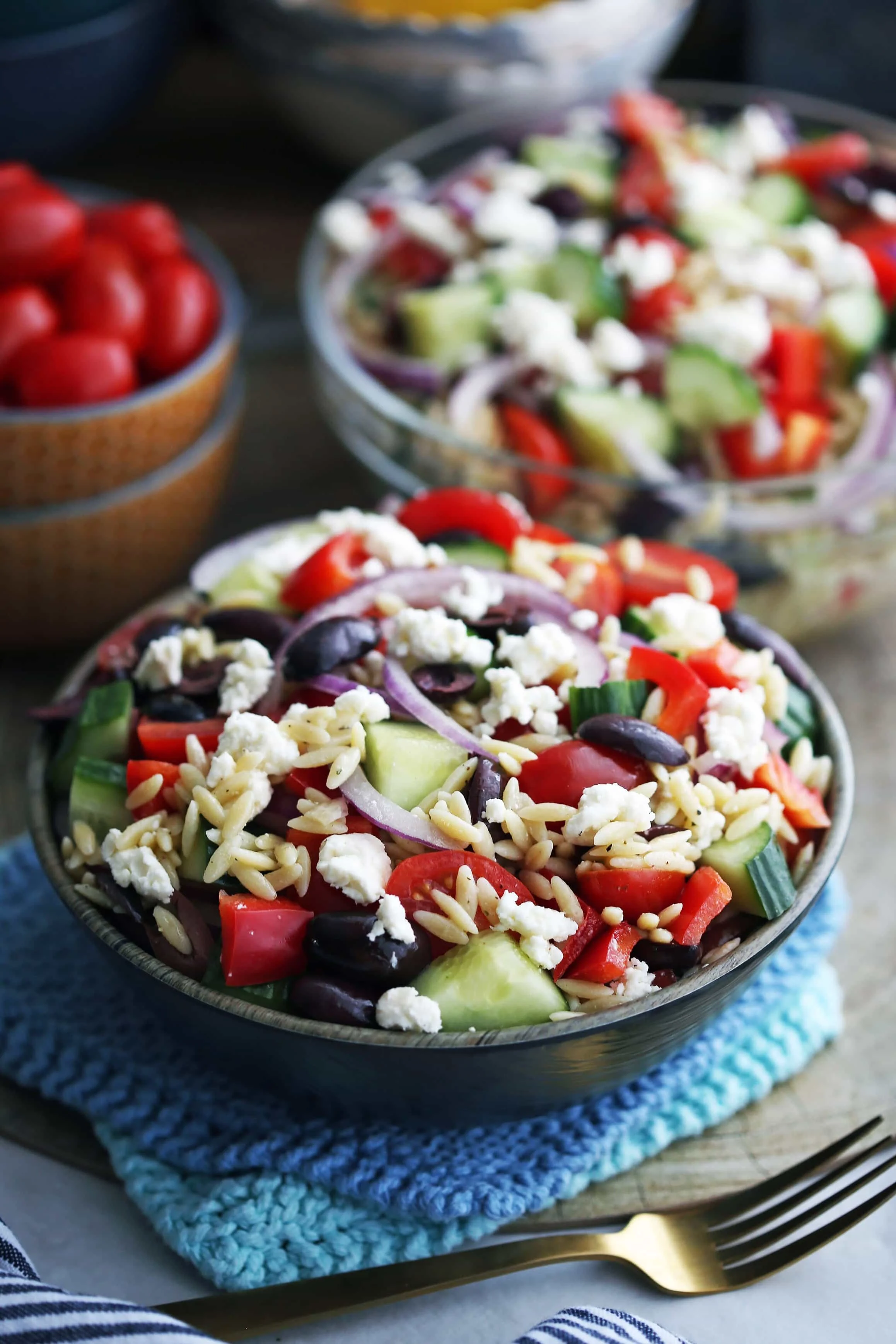 Greek orzo salad with crumbled feta, Kalamata olives, and raw vegetables in a dark brown bowl and a large glass bowl.