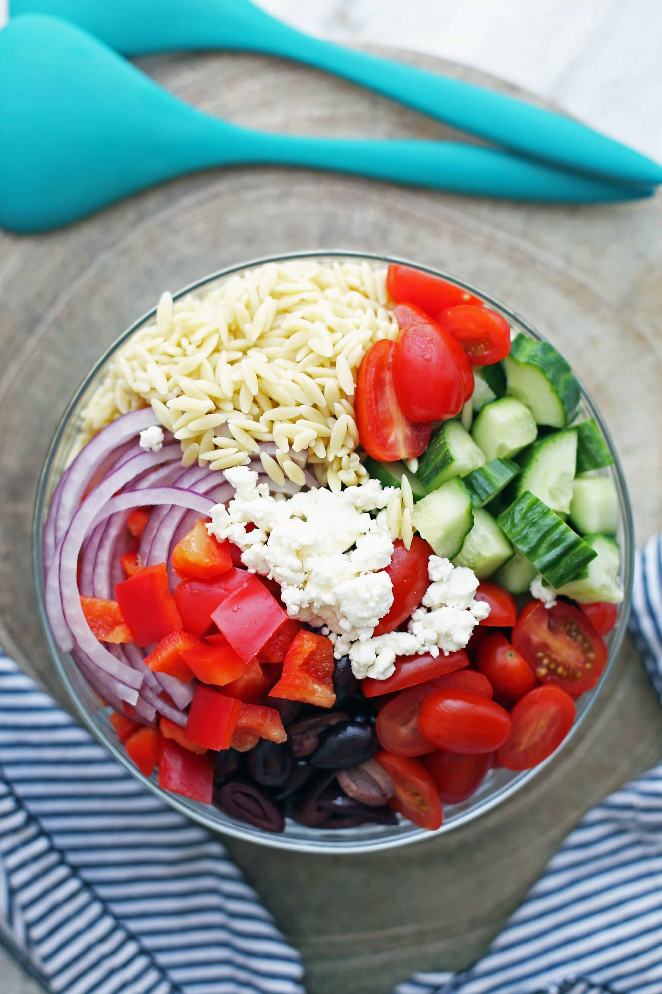 A large glass bowl containing orzo pasta, feta cheese, sliced red onions, and chopped tomatoes, olives, cucumber, and bell pepper.