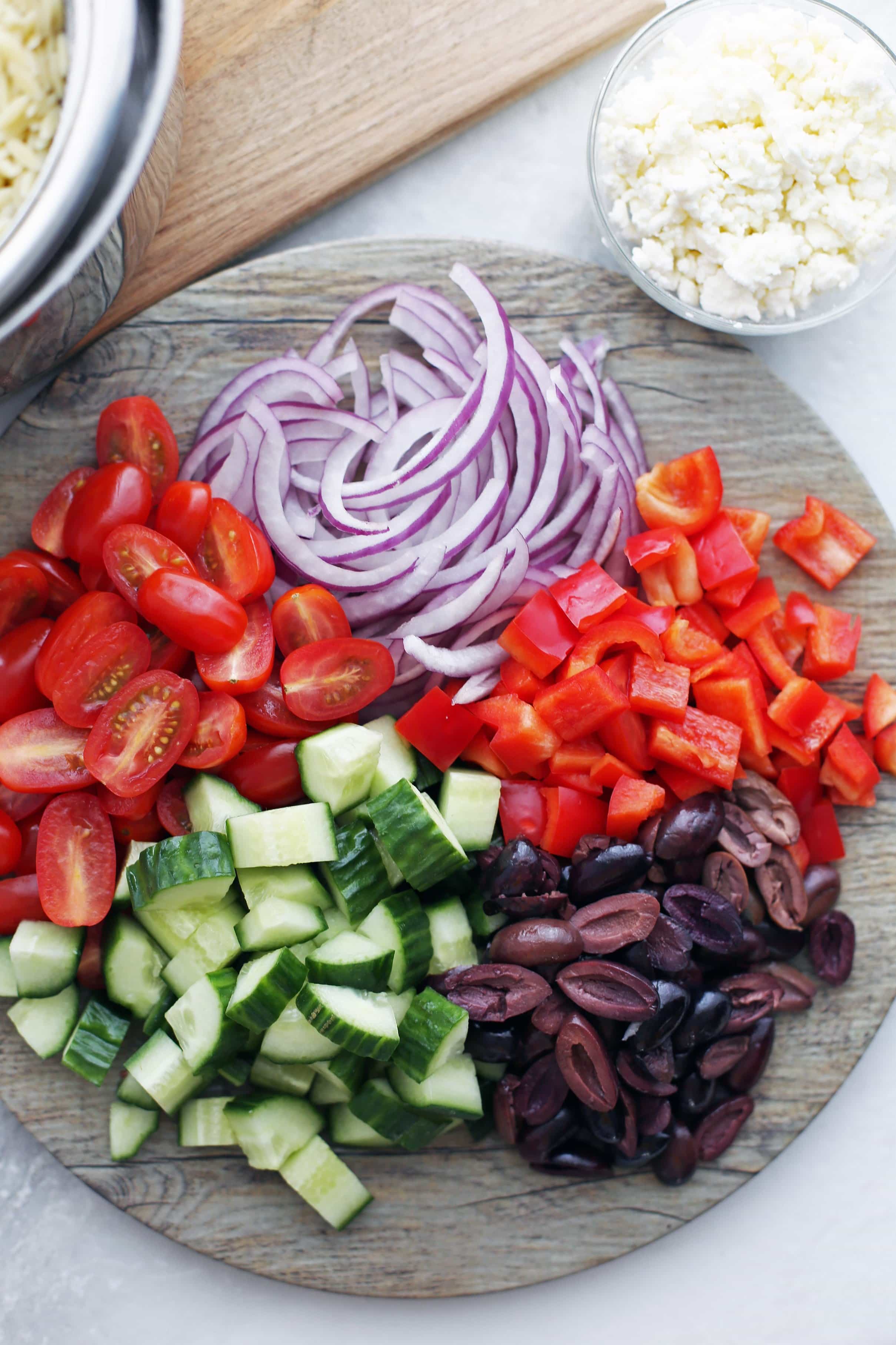 A large round wooden platter holding chopped cucumbers, grape tomatoes, red bell peppers, black olives, and red onions.