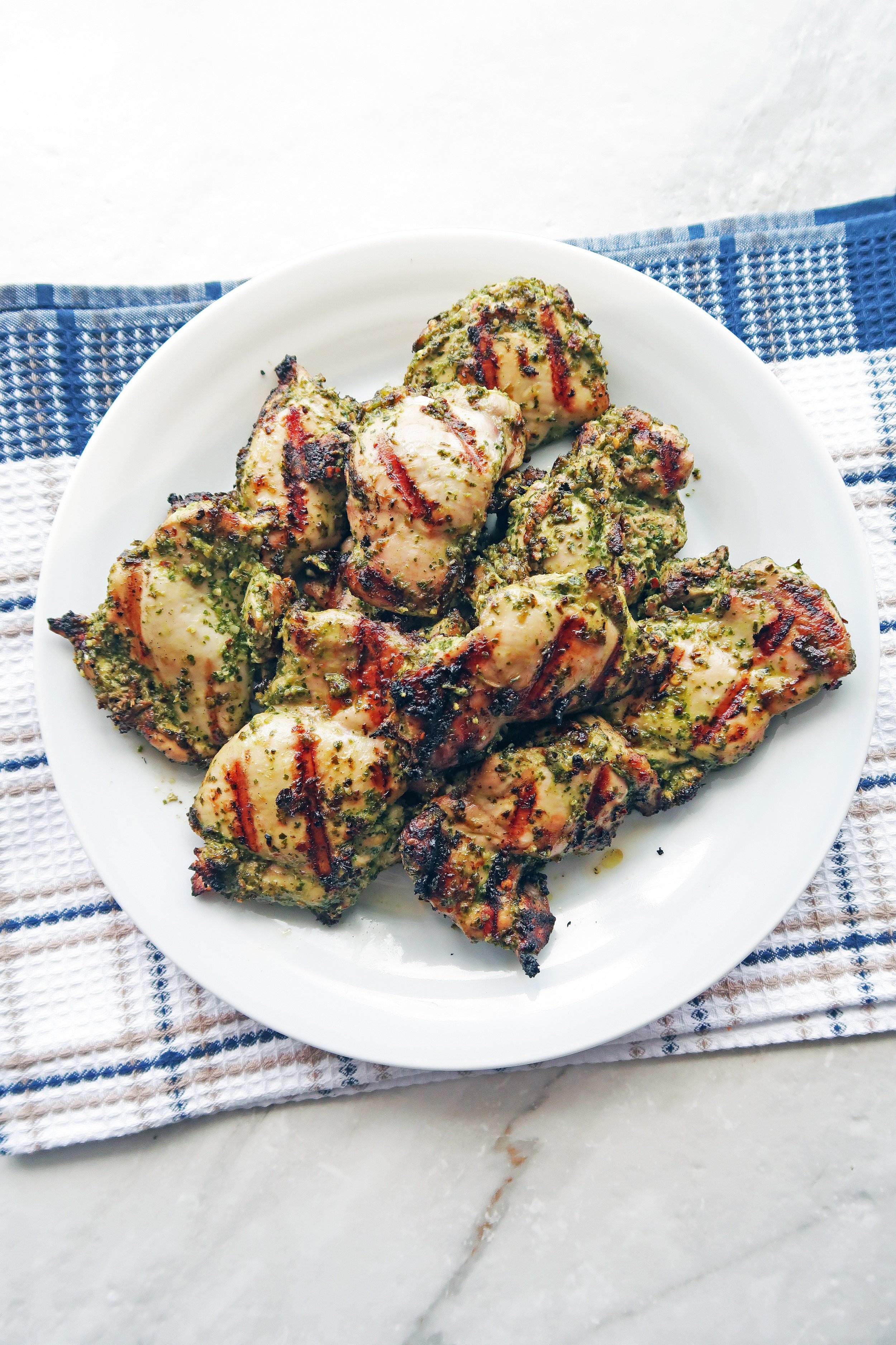 Overhead view of a white plate Grilled Chicken Thighs with Chimichurri Sauce on top of a blue checkered towel.