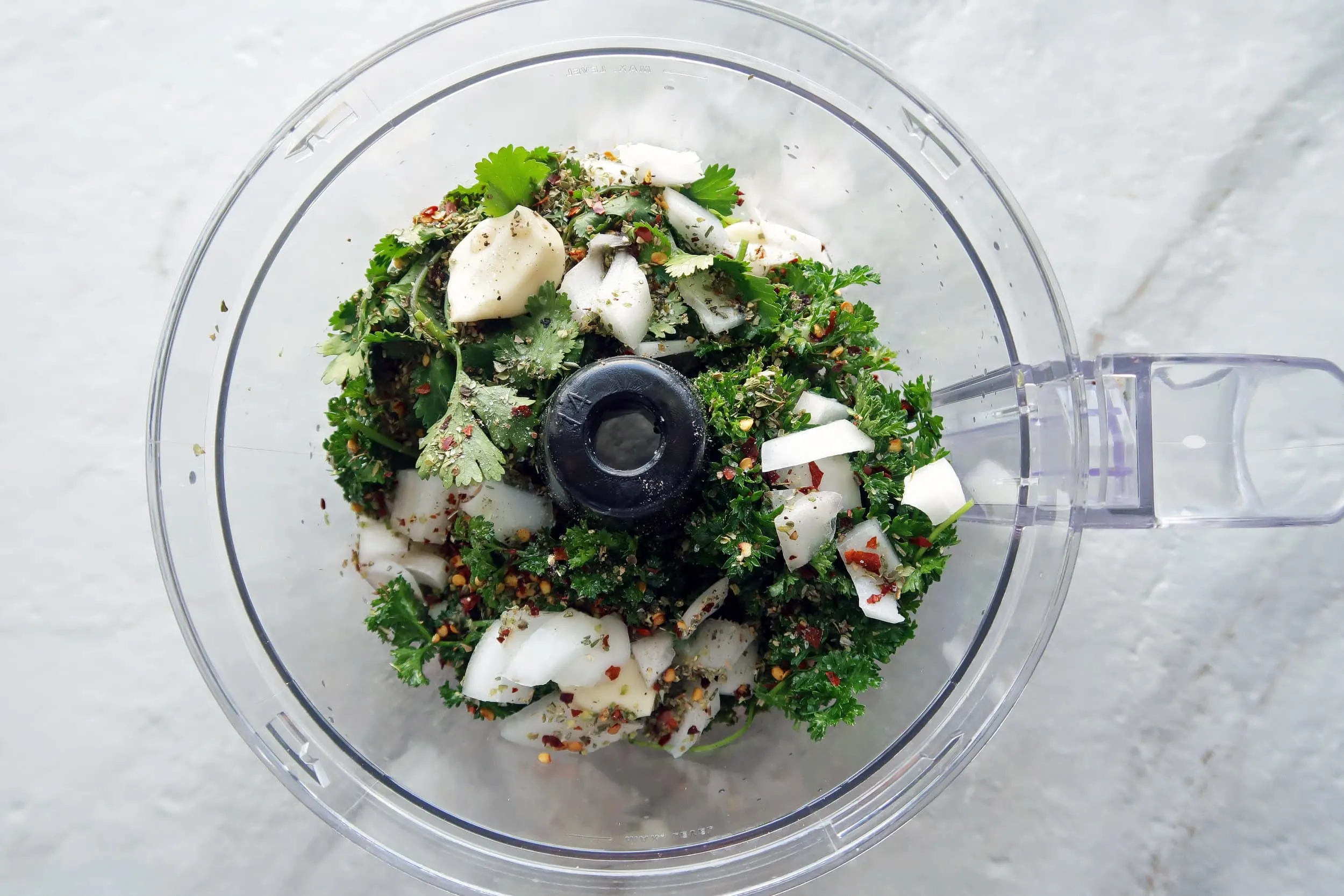 A food processor containing chimichurri sauce ingredients including parsley, garlic, and onion.