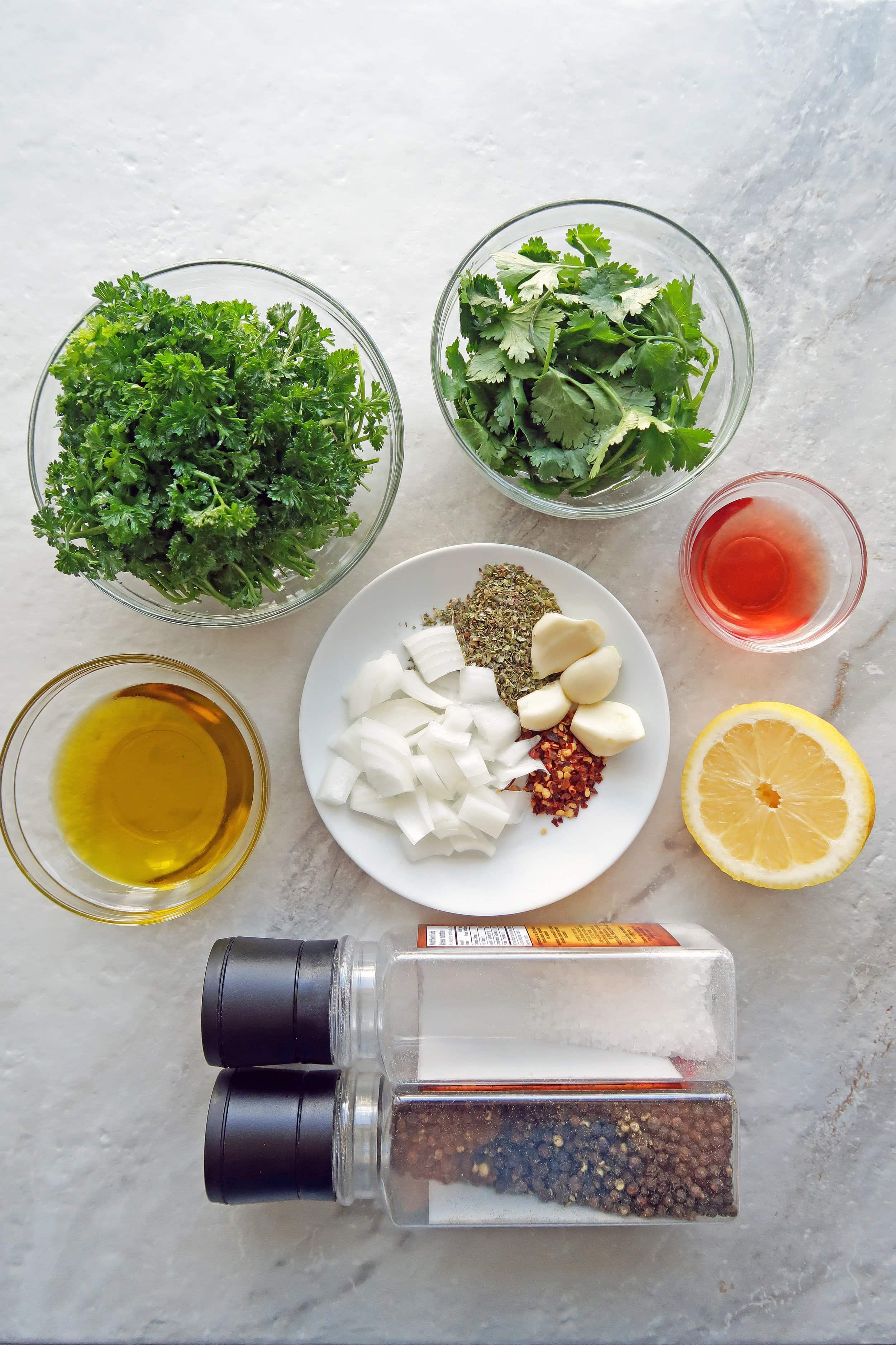 Bowls of parsley, cilantro, olive oil, and red wine vinegar alongside a lemon, chopped onions, garlic, oregano, and red pepper flakes.