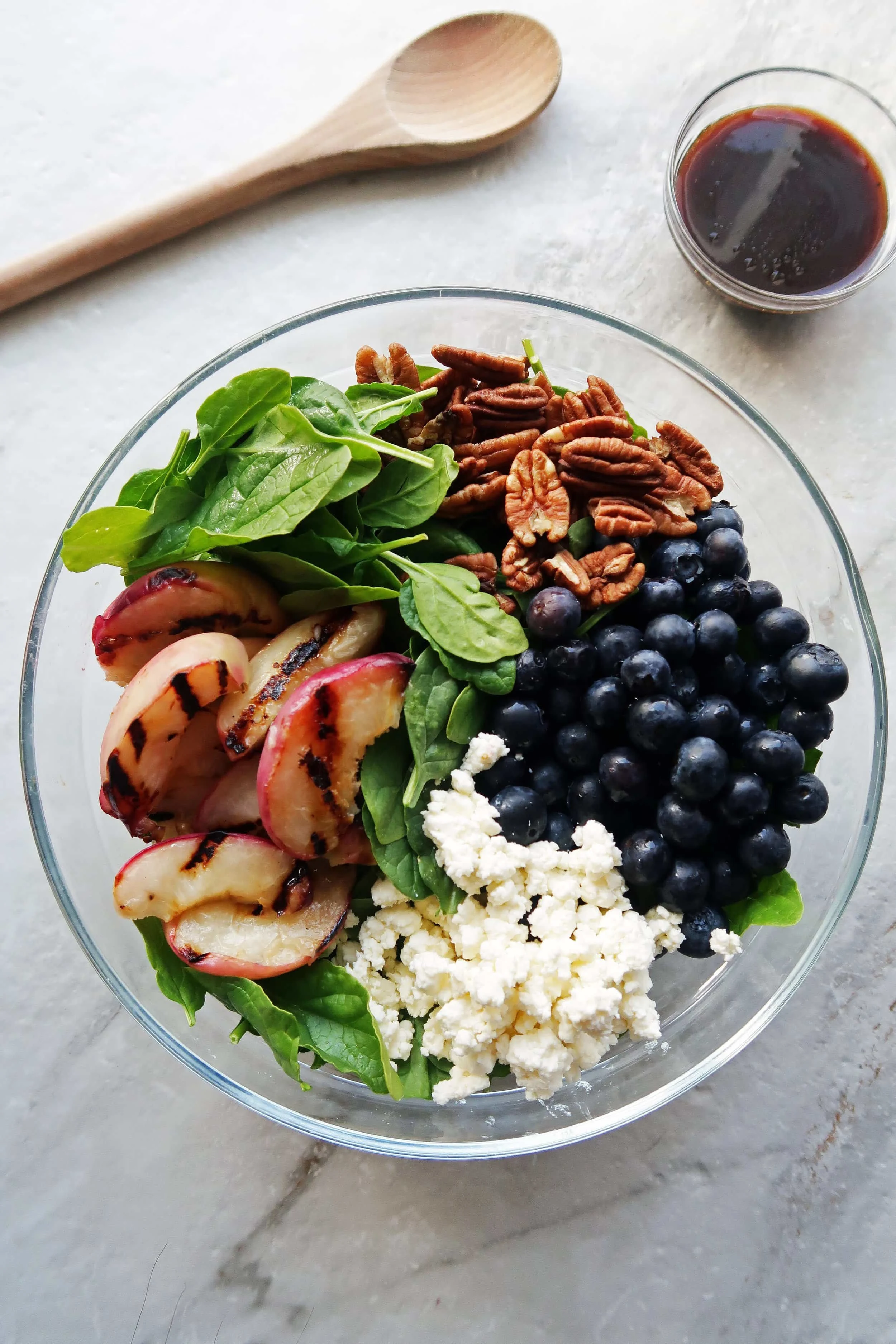A big glass bowl of spinach covered with grilled peach slices, pecans, blueberries, and crumbled feta cheese with honey balsamic vinaigrette on the side.
