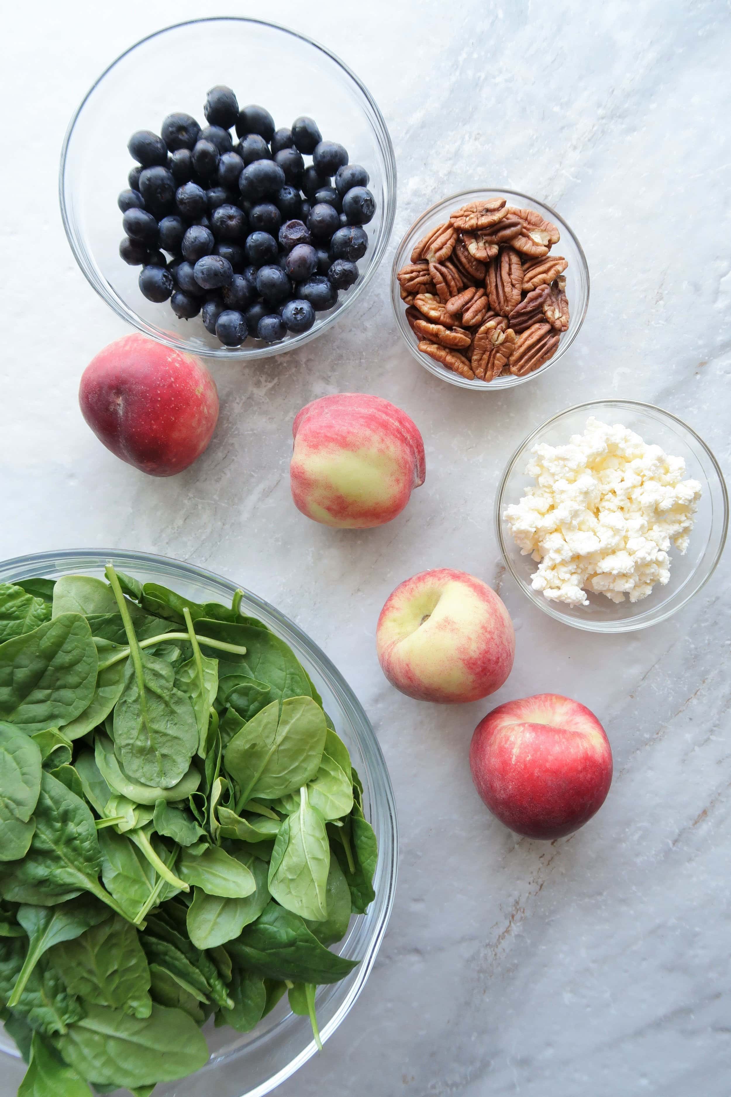 Glass bowls containing fresh blueberries, pecans,feta cheese, and spinach with four white peaches surrounding the bowls.