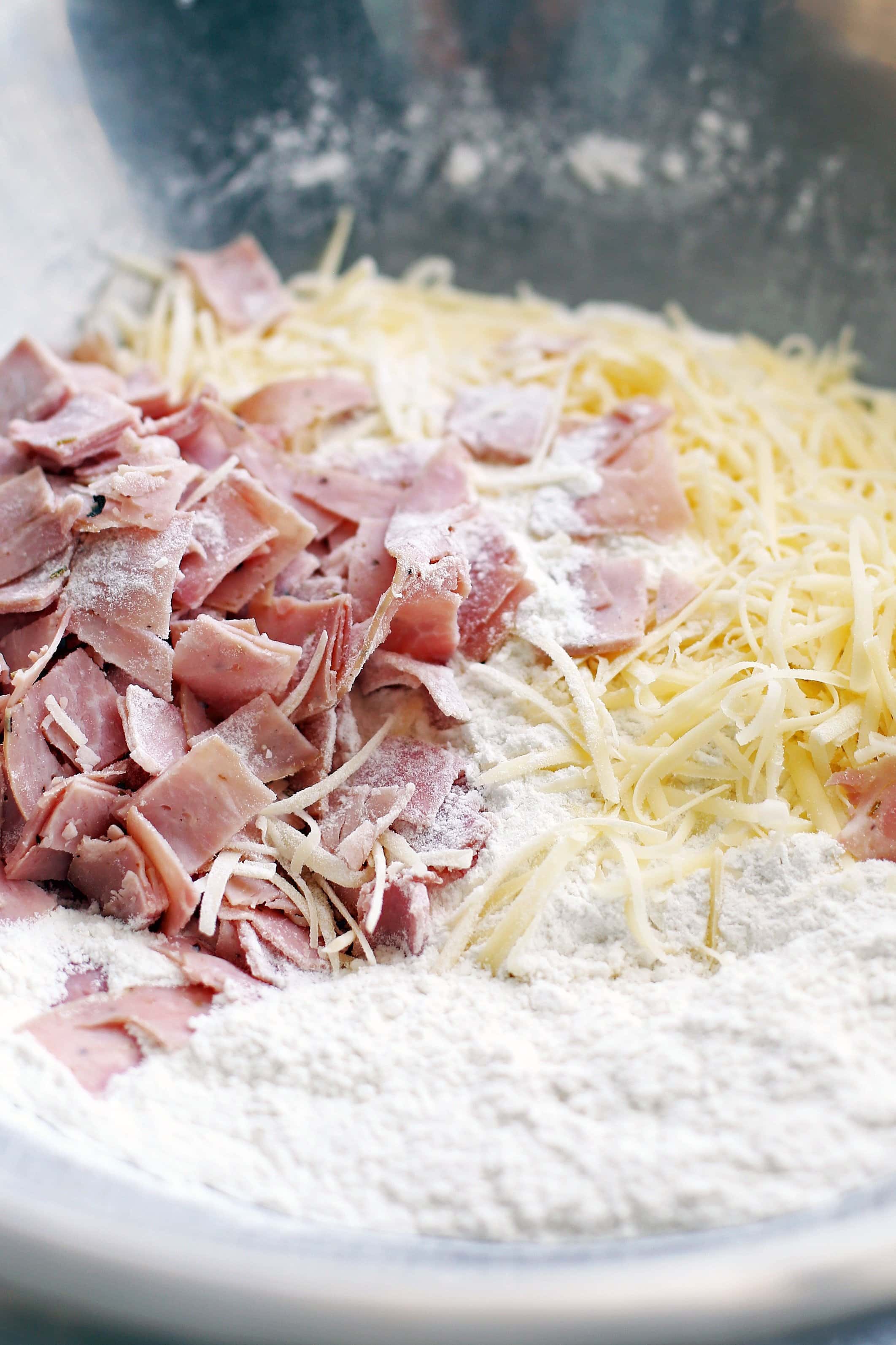 All-purpose flour, baking powder, sugar, salt, shredded cheese, and diced deli ham in a large stainless steel bowl.