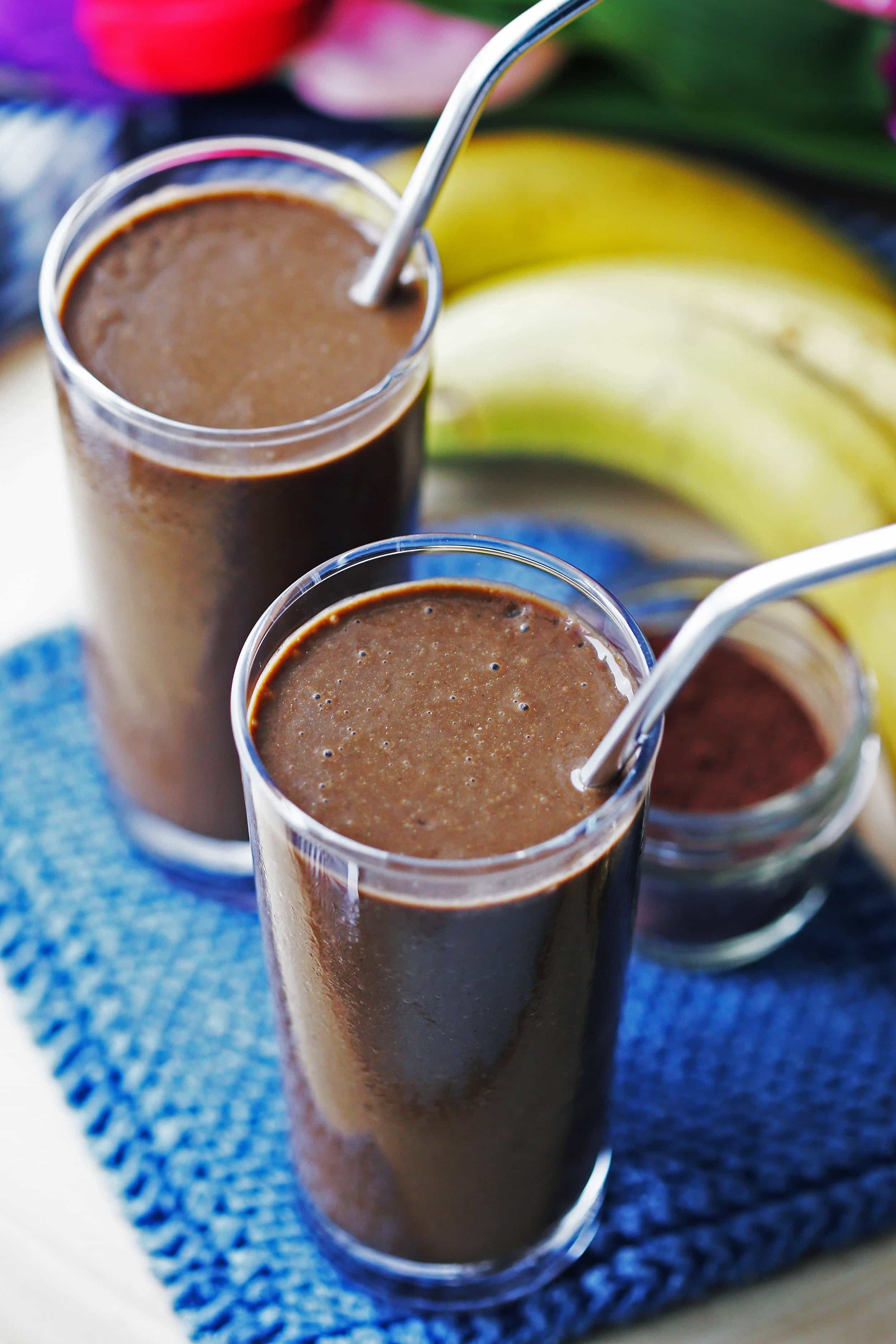 Healthy chocolate banana smoothies in two tall glasses with bananas and a small jar of cocoa powder behind them.