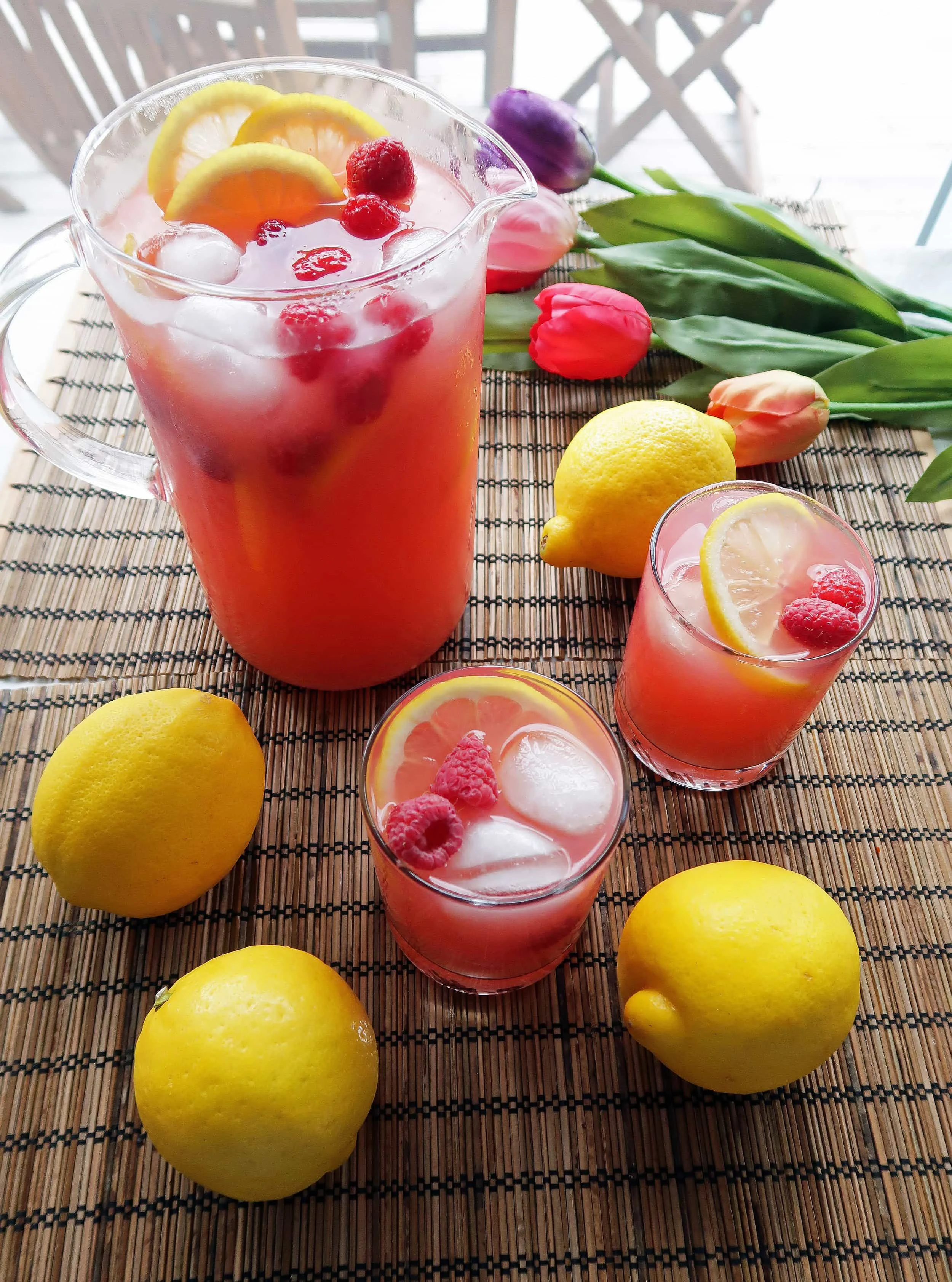 Two glasses and a pitcher full of homemade Raspberry Green Tea Lemonade with lemon slices and raspberry garnish; whole lemons around them.