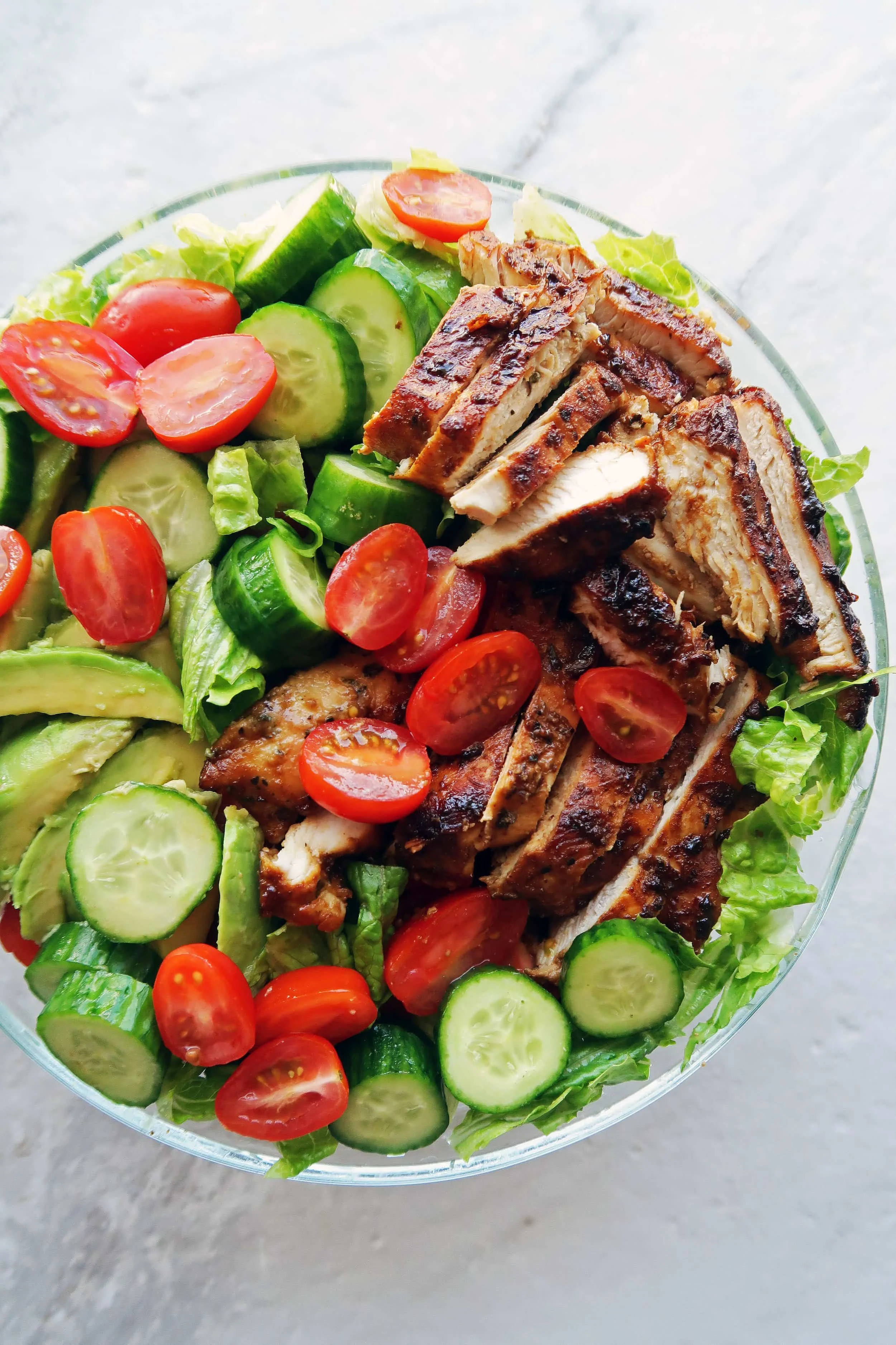 Chopped lettuce, tomatoes, cucumbers, avocado, and sliced honey mustard chicken breasts in a large bowl.