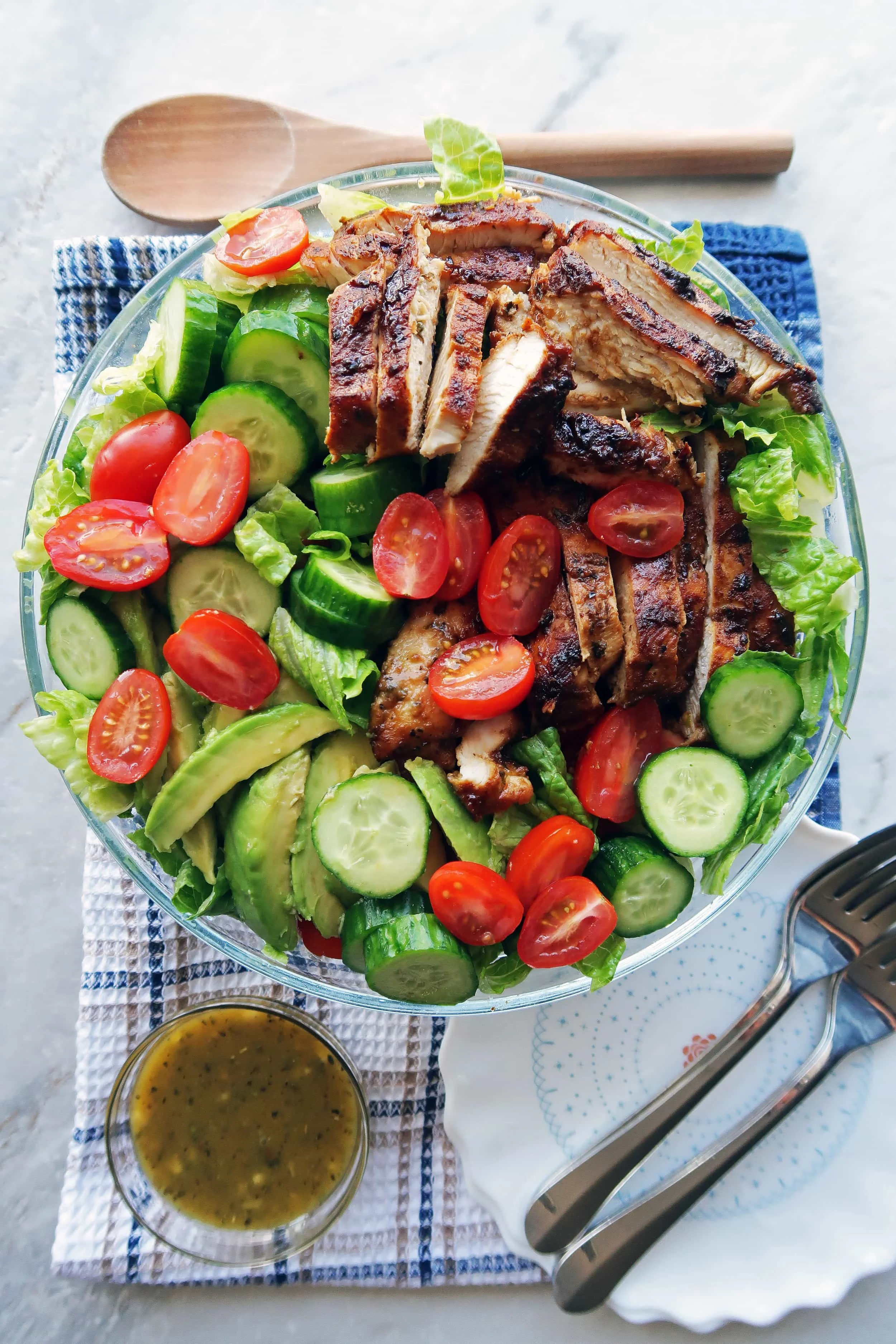 Lettuce, tomatoes, cucumbers, avocado, and sliced honey mustard chicken in a large bowl with dressing on the side.