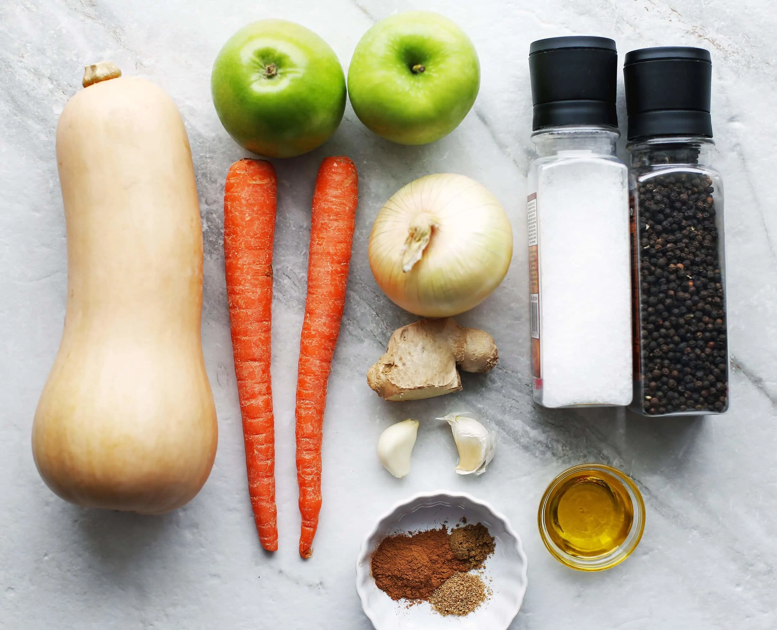 An overshot shot of fresh butternut squash, carrots, onion, ginger, garlic, salt, pepper, spices, and olive oil.